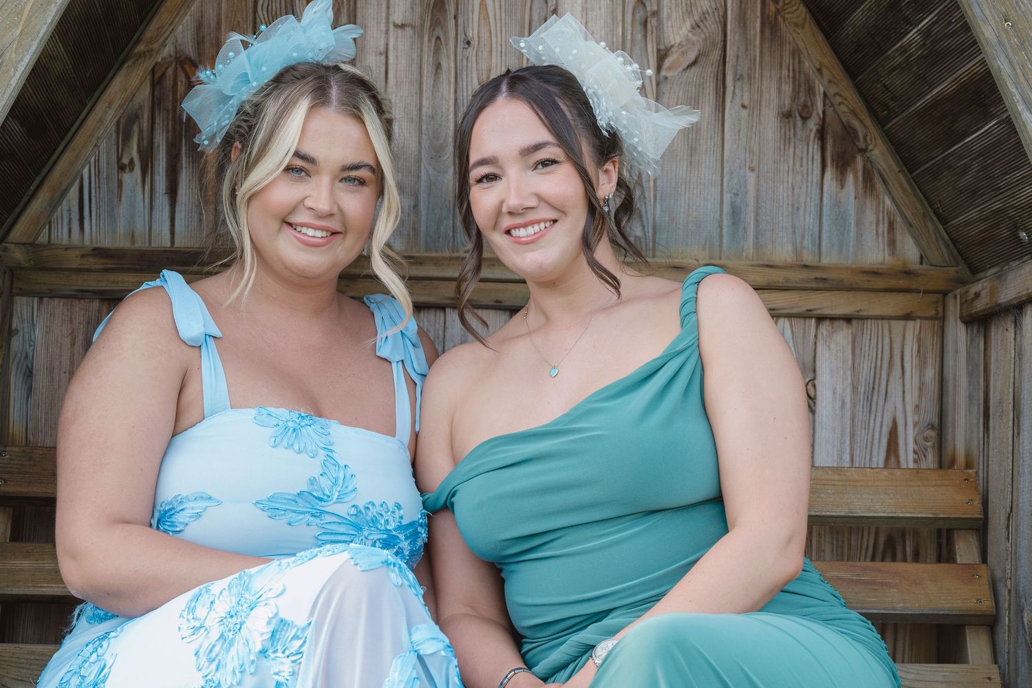 Two women in blue formal dresses and floral headpieces posing together at an outdoor event.