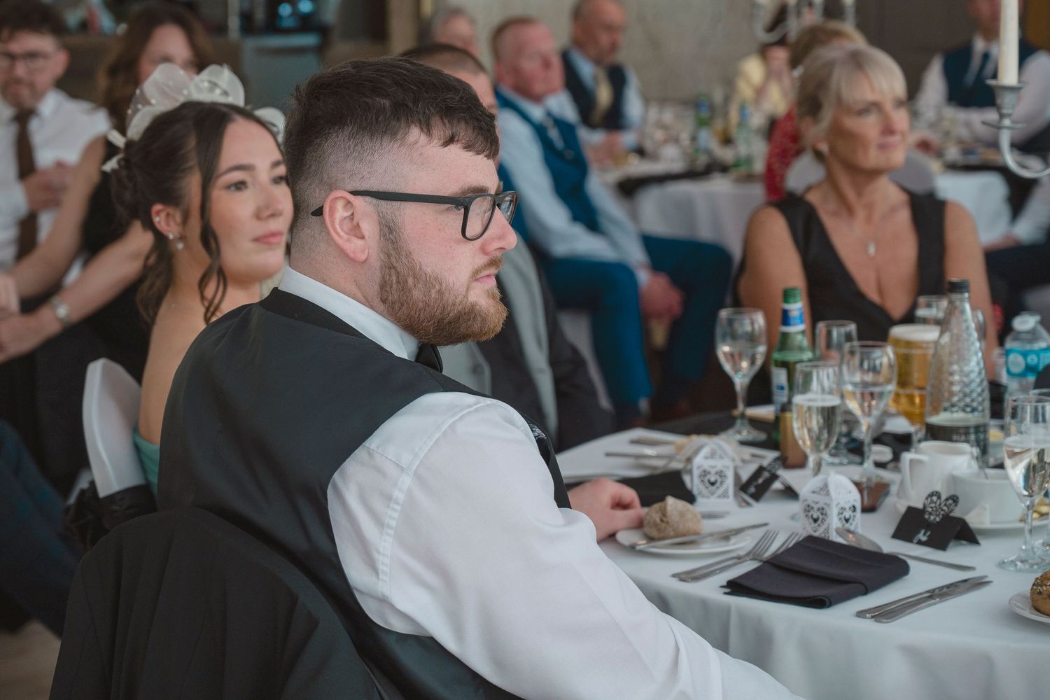 Guests seated at a wedding reception dinner, man in glasses and vest in foreground, elegant venue setting.