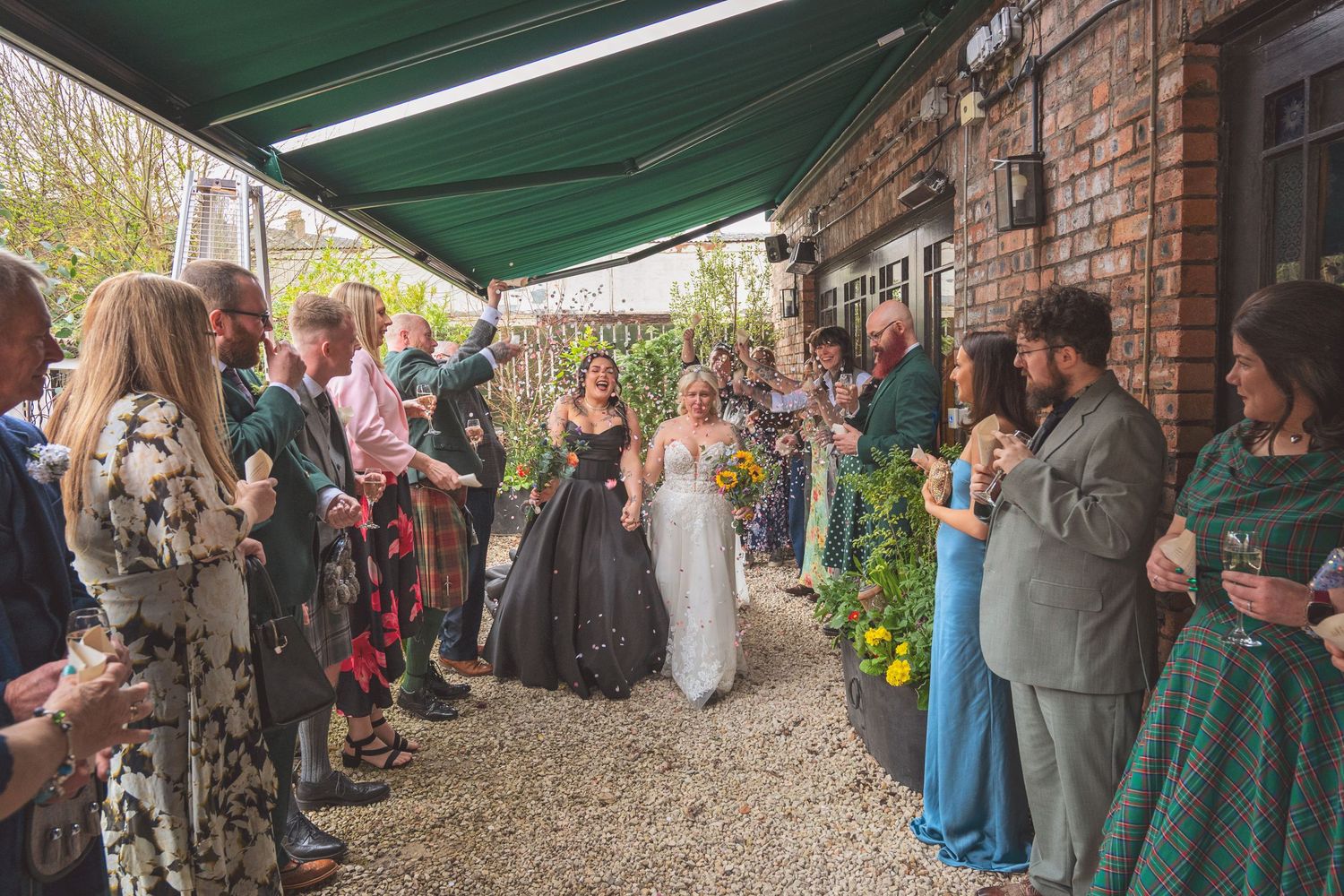 Two brides walk through a confetti tunnel of wedding guests outside a rustic venue.