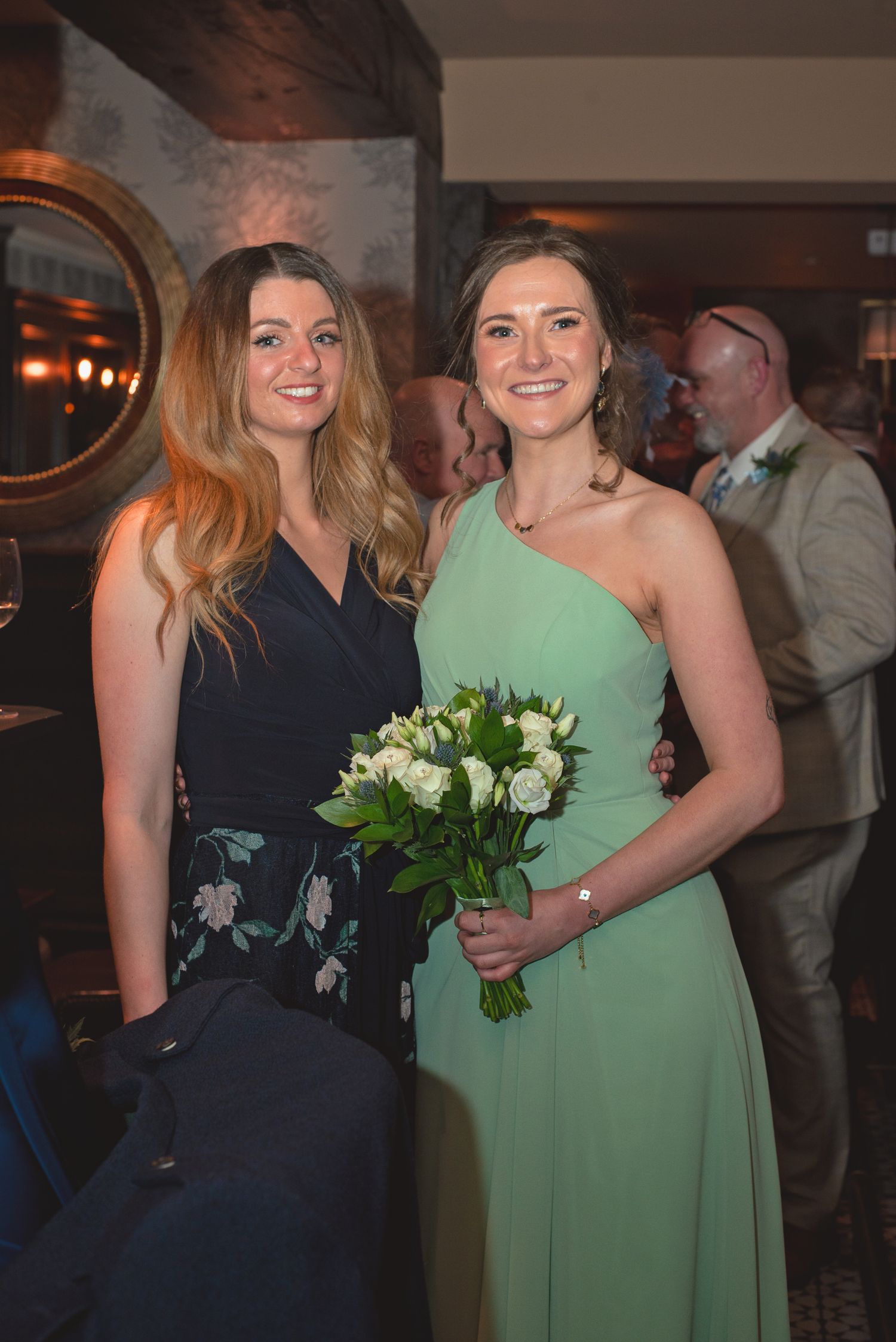 Two women in elegant dresses pose at a formal event, one holding a white floral bouquet.