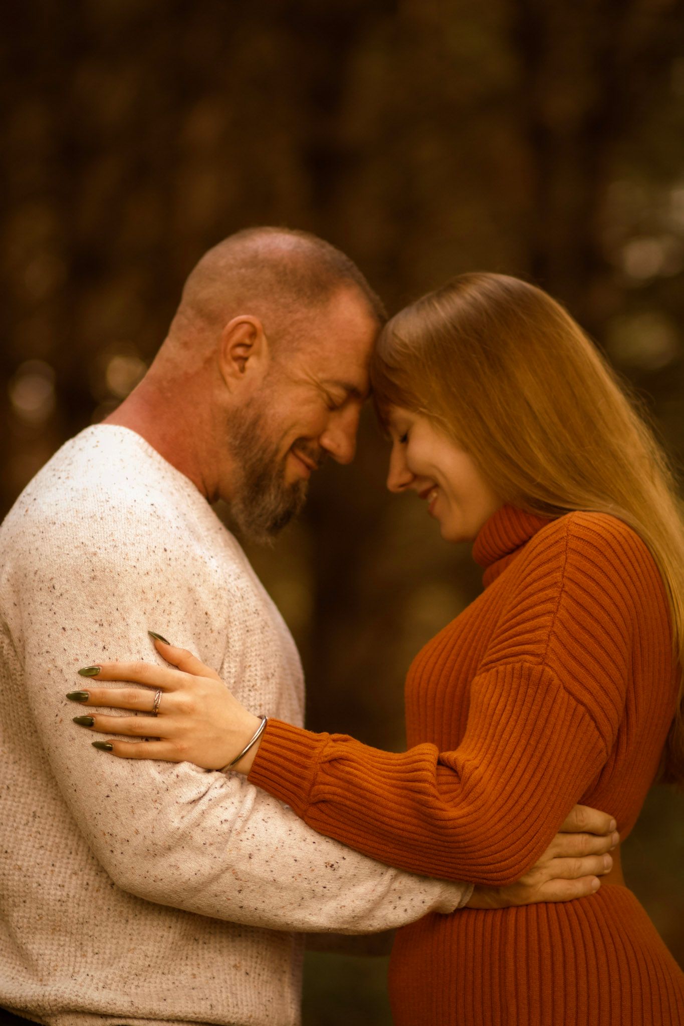 A smiling couple embraces tenderly in an autumn forest, foreheads touching, wearing cozy sweaters in warm fall colors.