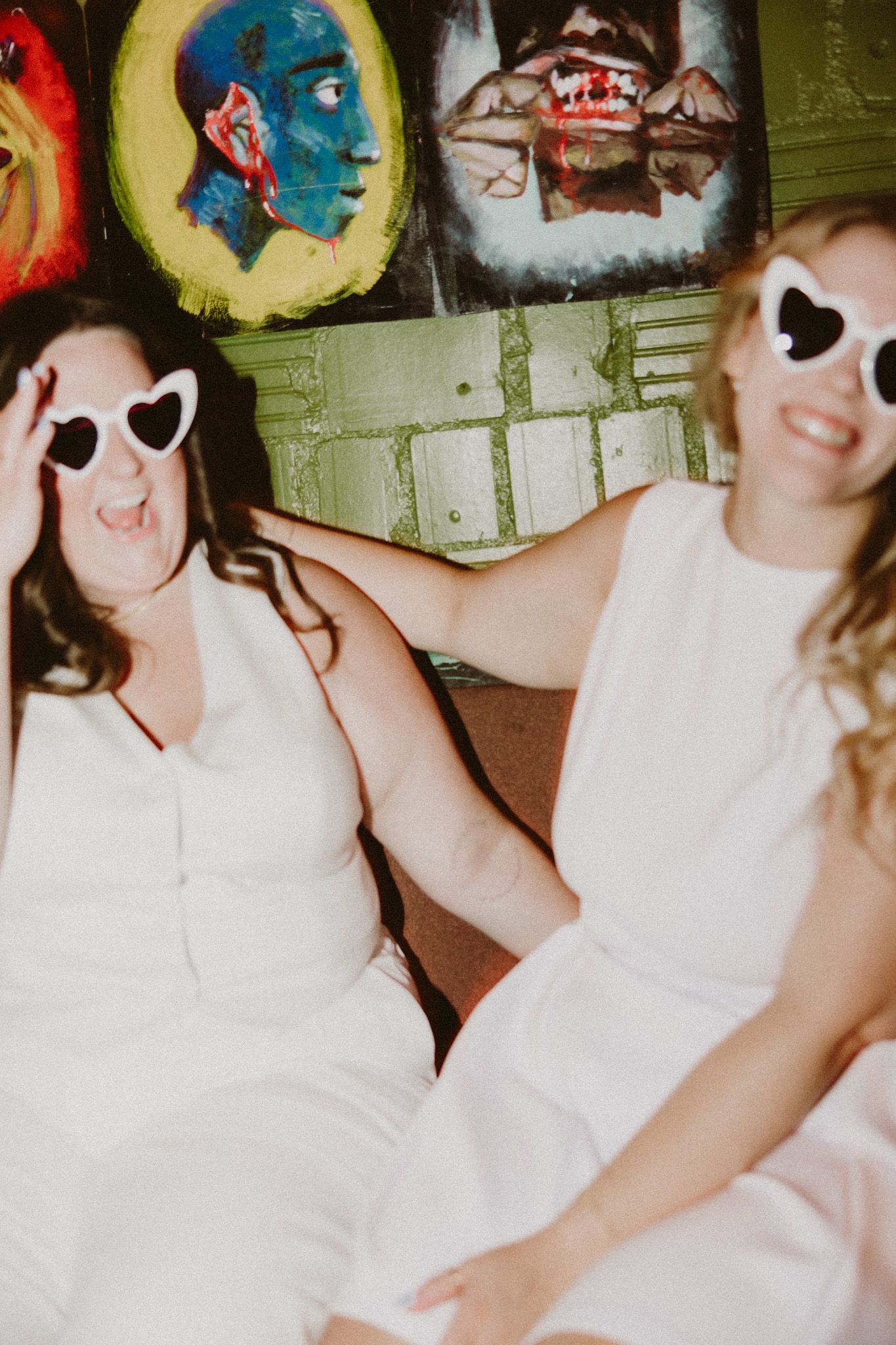Two women in white dresses wearing heart-shaped sunglasses sitting together in front of colorful artwork paintings.