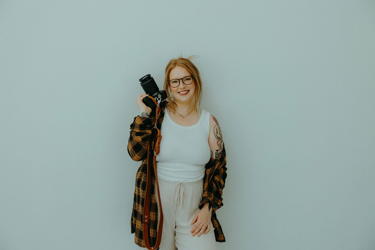 Smiling redhead woman with glasses holding a camera, wearing a plaid shirt and white tank top against a light blue wall.