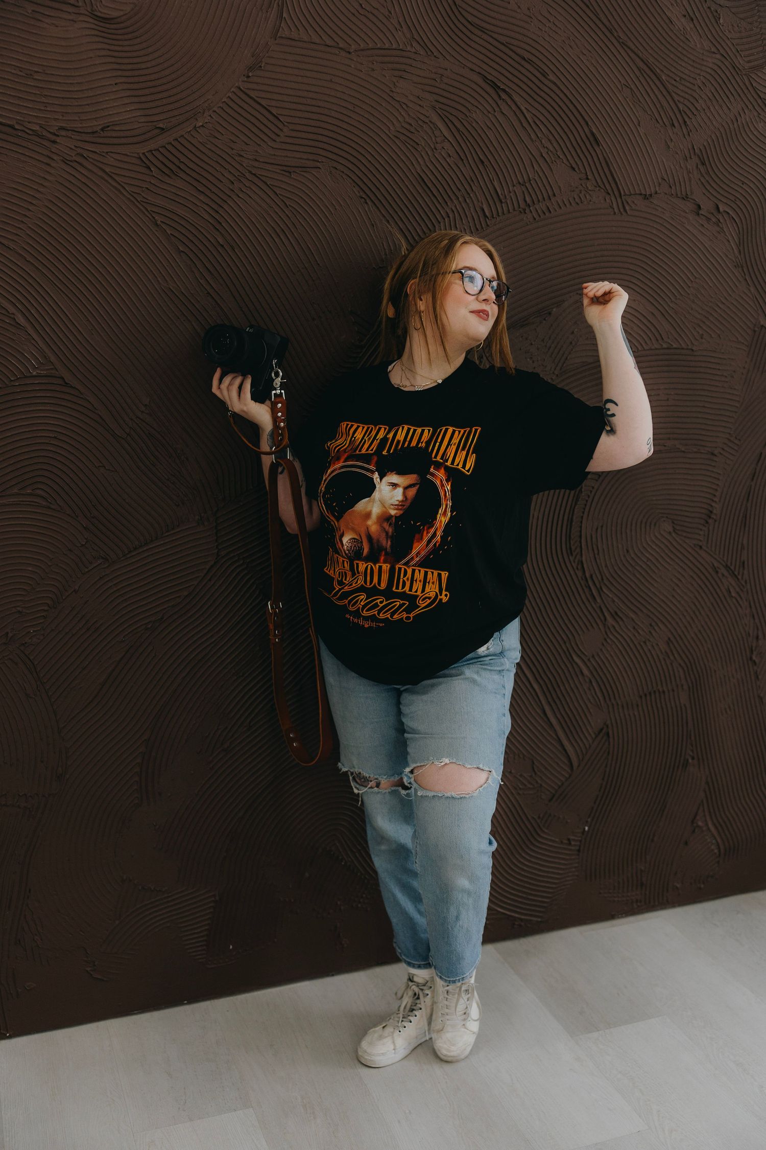 Woman in black graphic tee, ripped jeans, and white sneakers posing confidently against a textured dark wall.