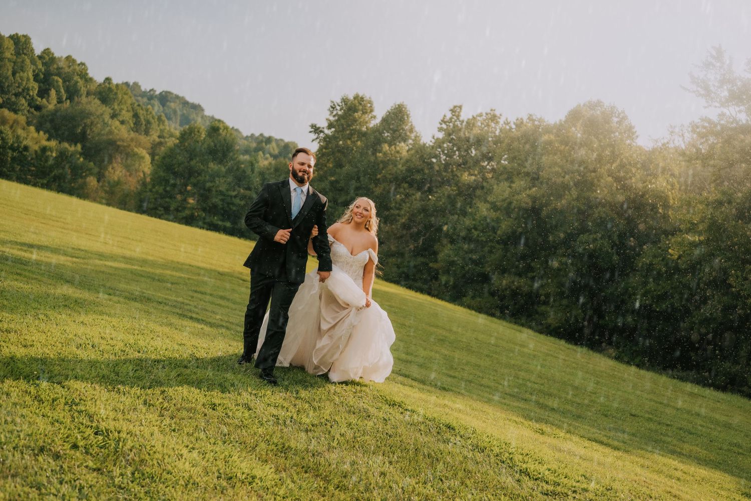 Bride and groom running through rain on lush green hillside at golden hour wedding celebration.