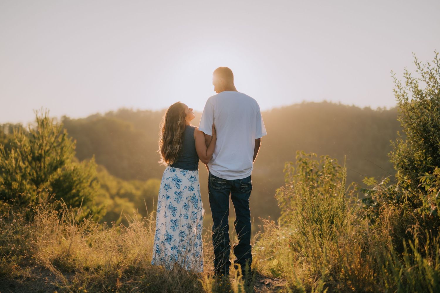 Couple stands together in a golden field at sunset with mountains in the background.