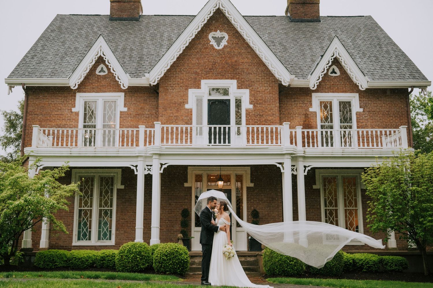 A romantic wedding moment outside a beautiful Victorian-style brick mansion with white trim and wraparound porch.