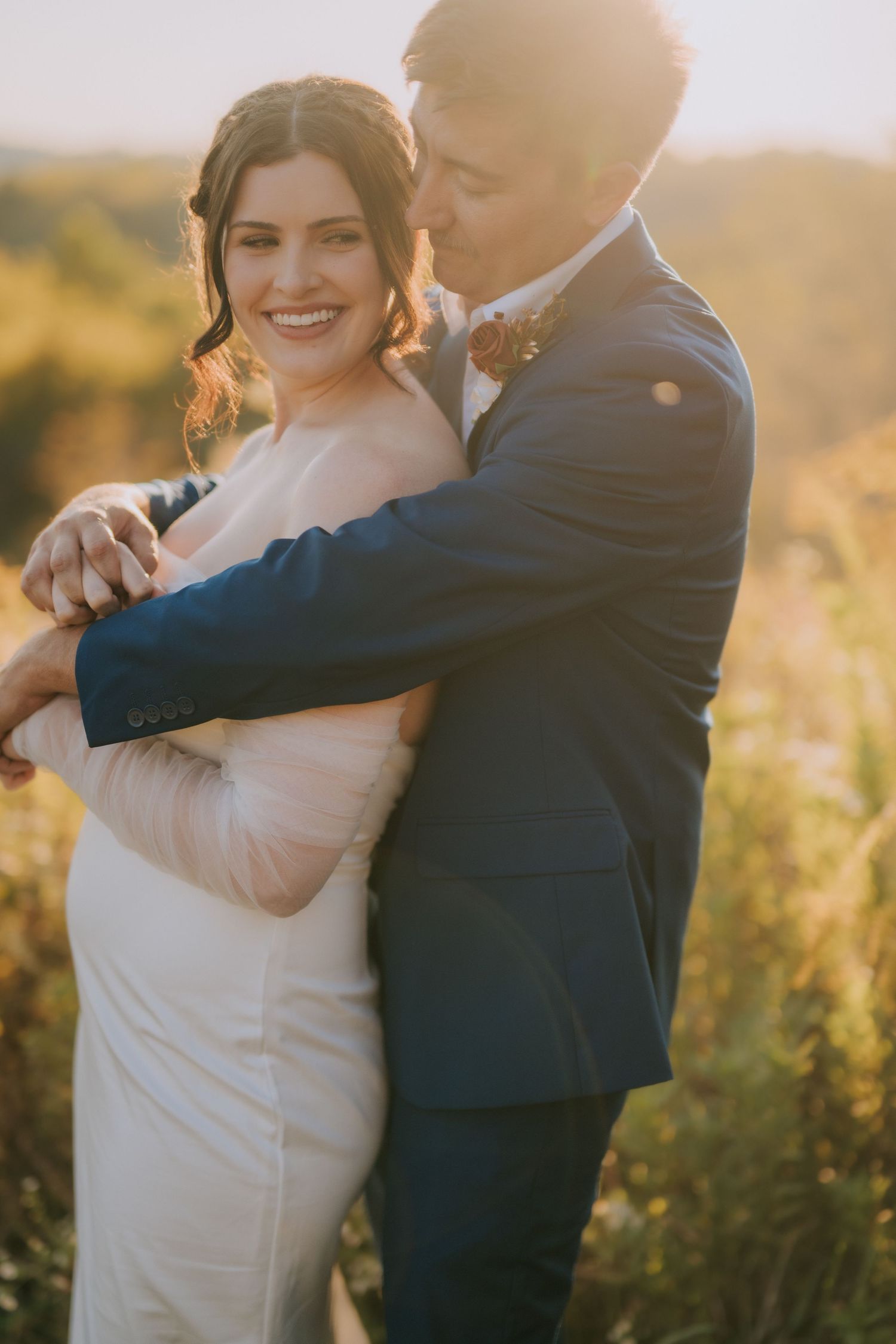 A romantic wedding couple embraces in a golden field at sunset during their outdoor photo session.