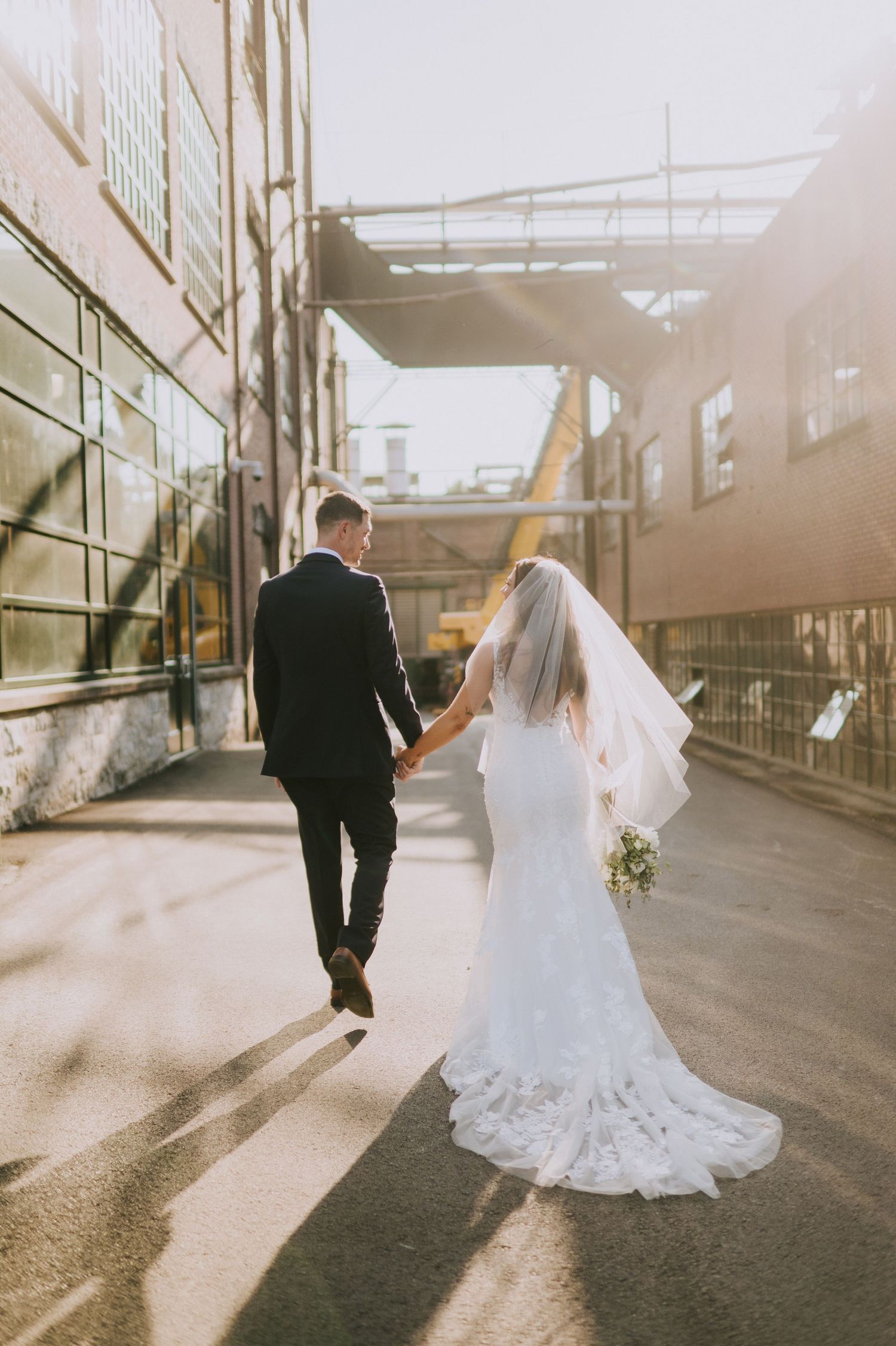 Couple in wedding attire walking down an industrial corridor in golden sunlight.