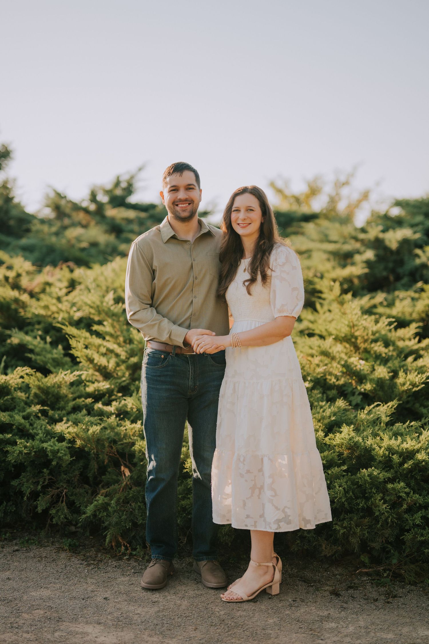 A couple poses together against a backdrop of green shrubs in casual formal attire.
