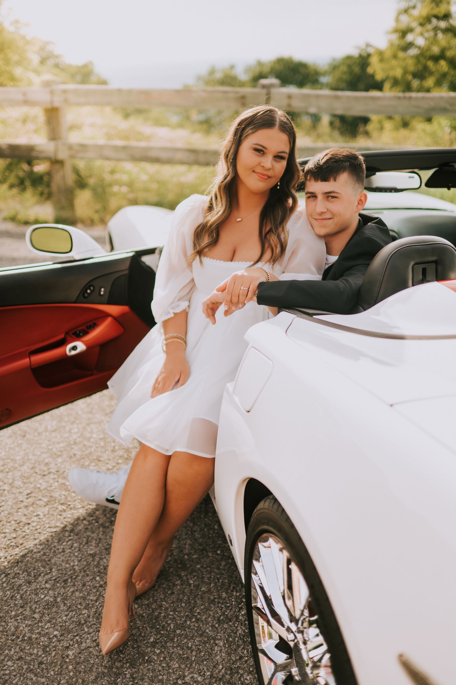 A stylish couple poses with a white sports car in warm sunset lighting.