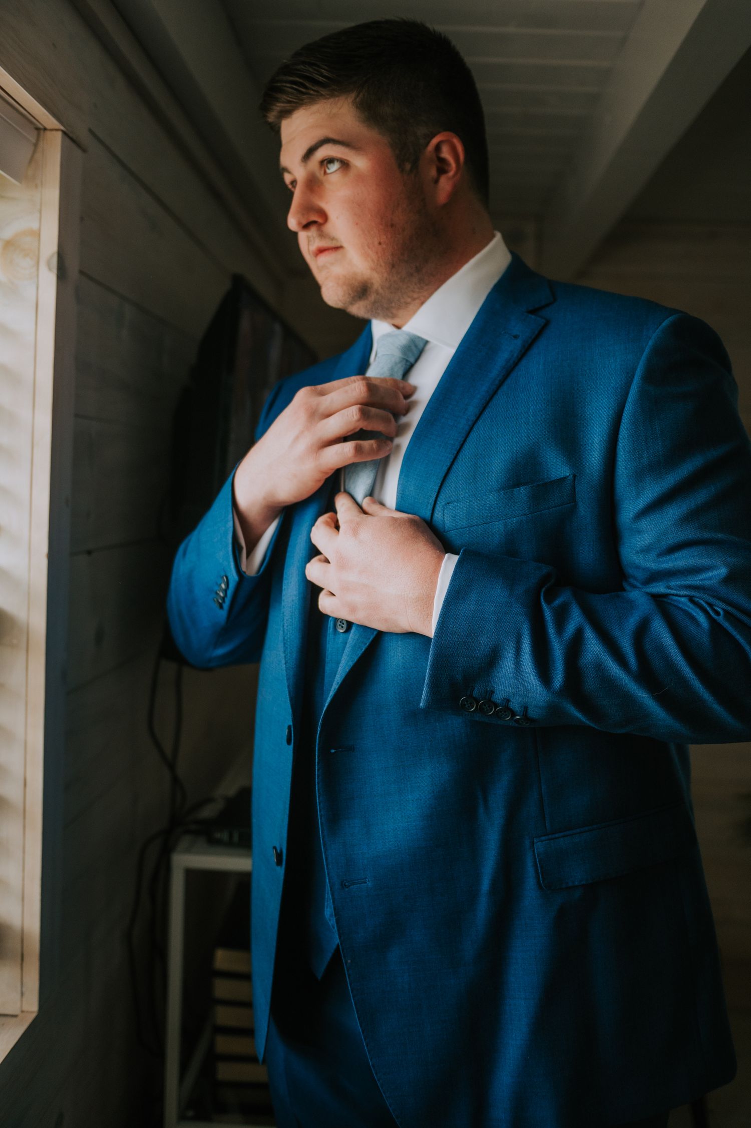 Man in navy blue suit adjusting tie in dramatic window lighting.