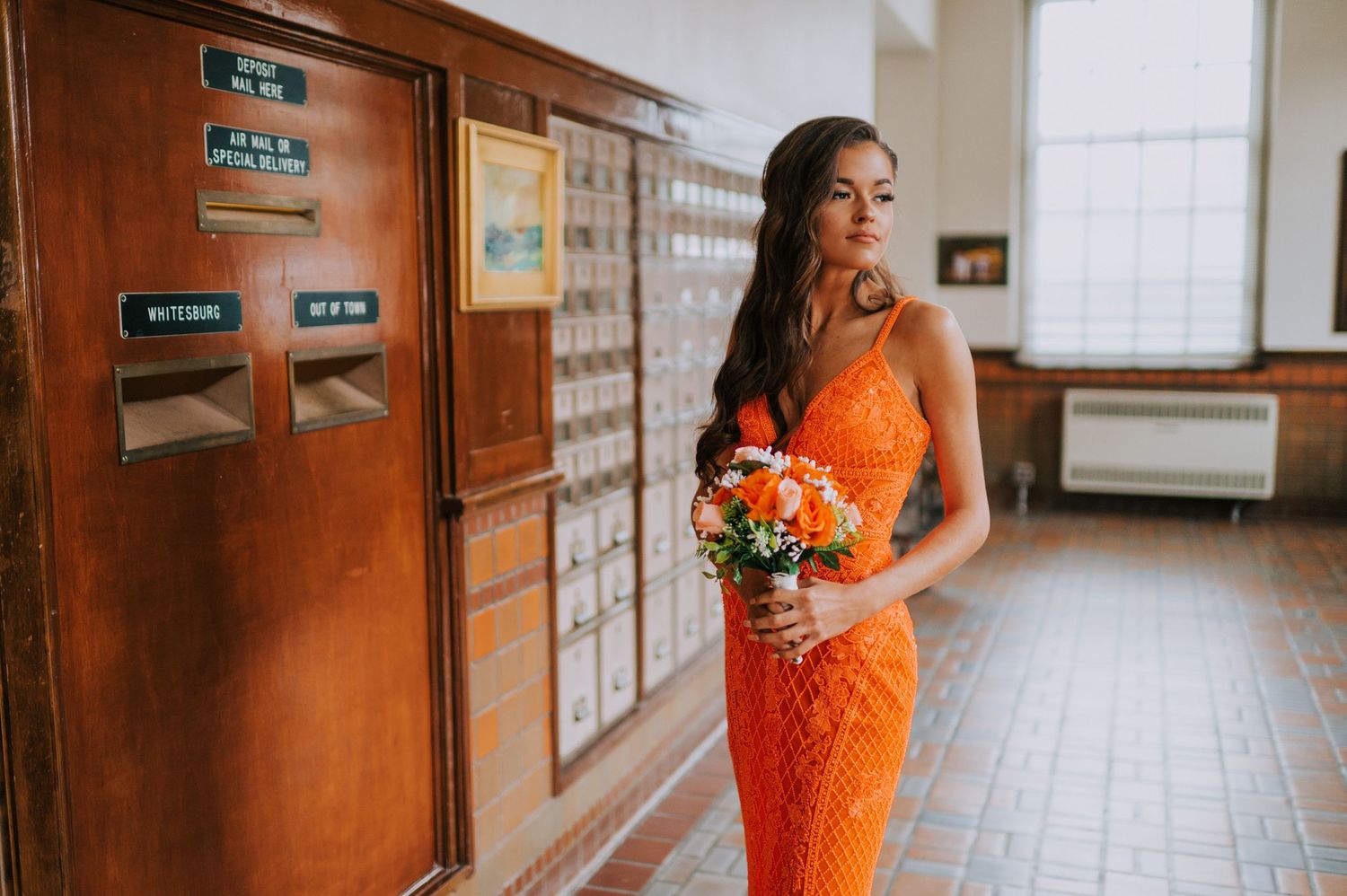 Model in bright orange dress poses in a hallway with wooden doors and brick walls.