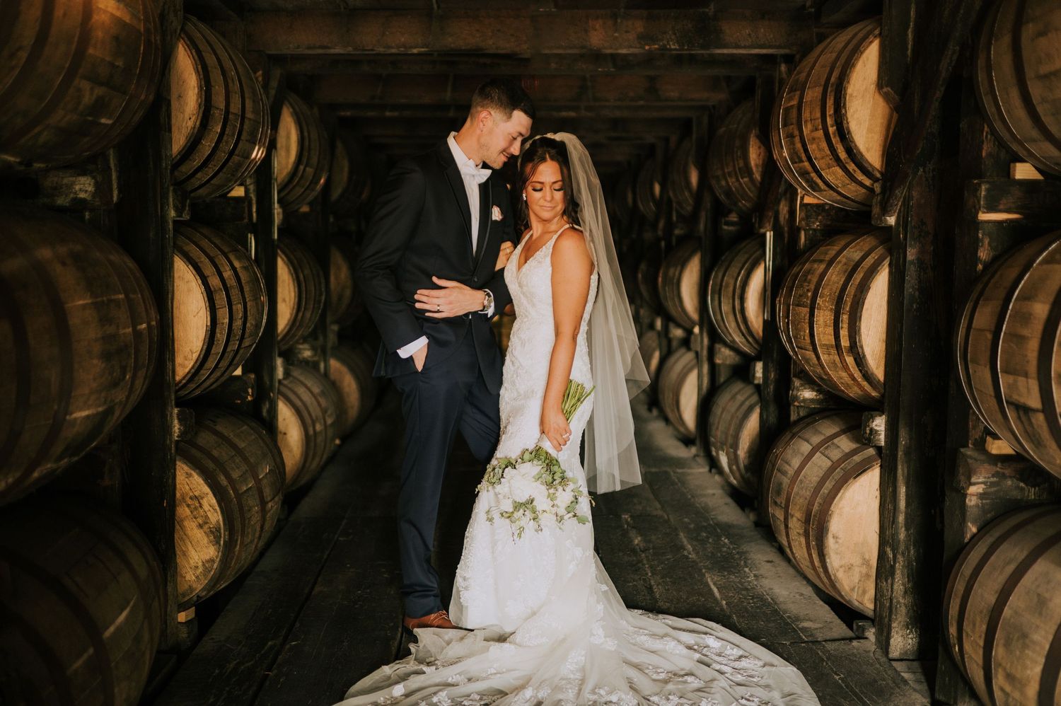 Rustic wedding photo session among wooden whiskey barrels in dimly lit venue.