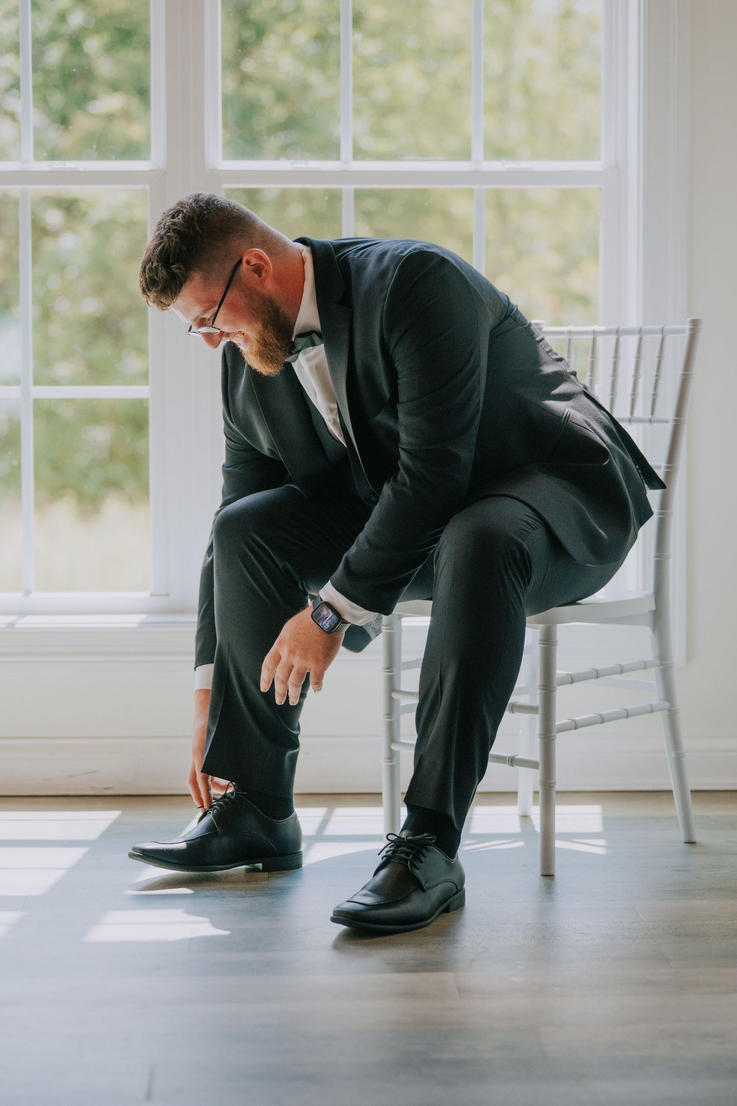 Groom checking his shoes in bright sunlit room with white chairs.