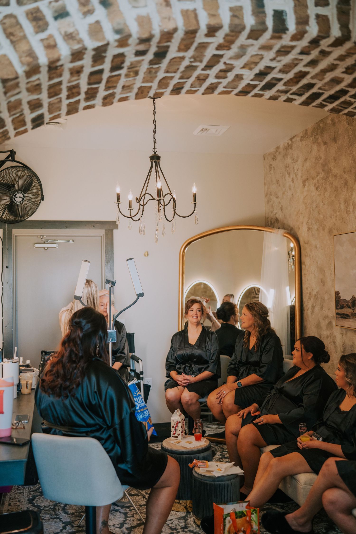 Wedding preparation scene in rustic brick room with chandelier and mirrors.