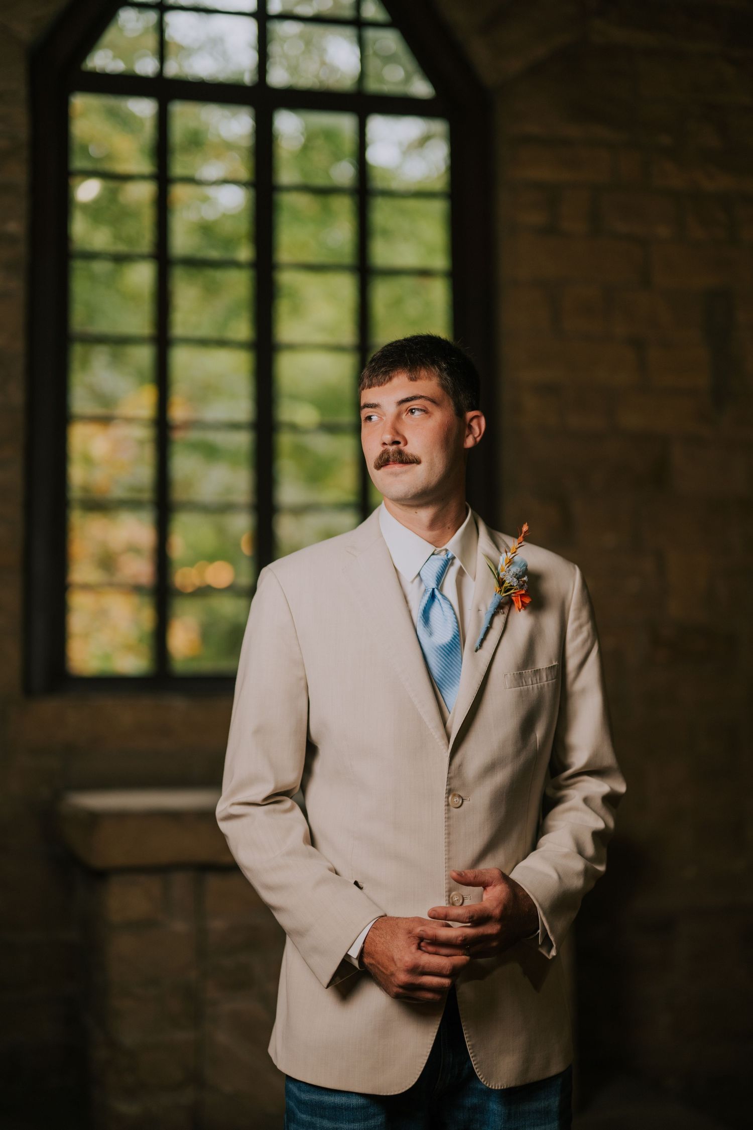 A person in a beige suit and light blue tie stands in front of stone walls and windows.