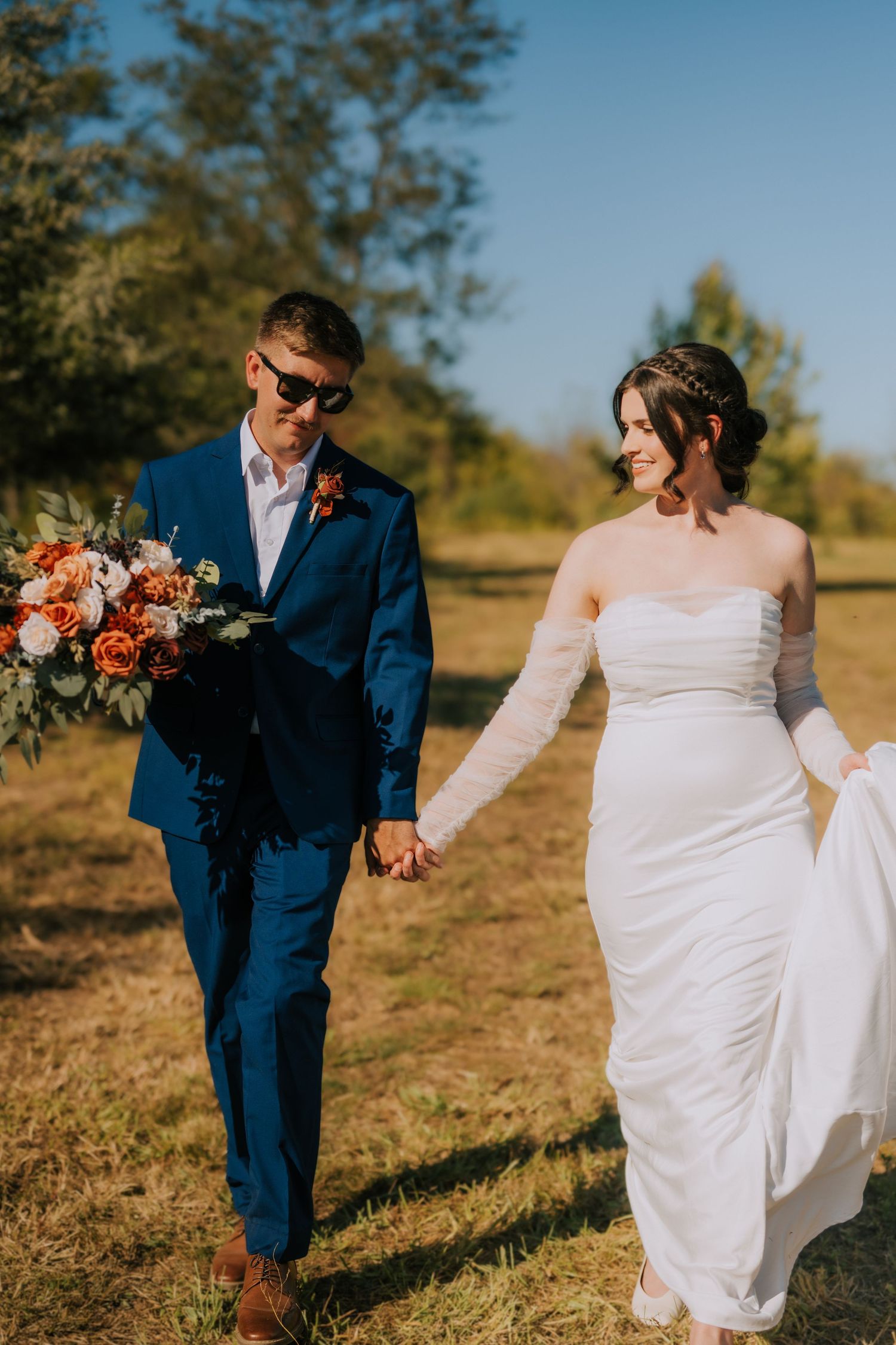 A wedding couple walks hand in hand through a sunny field, with bride in white and groom in navy blue.