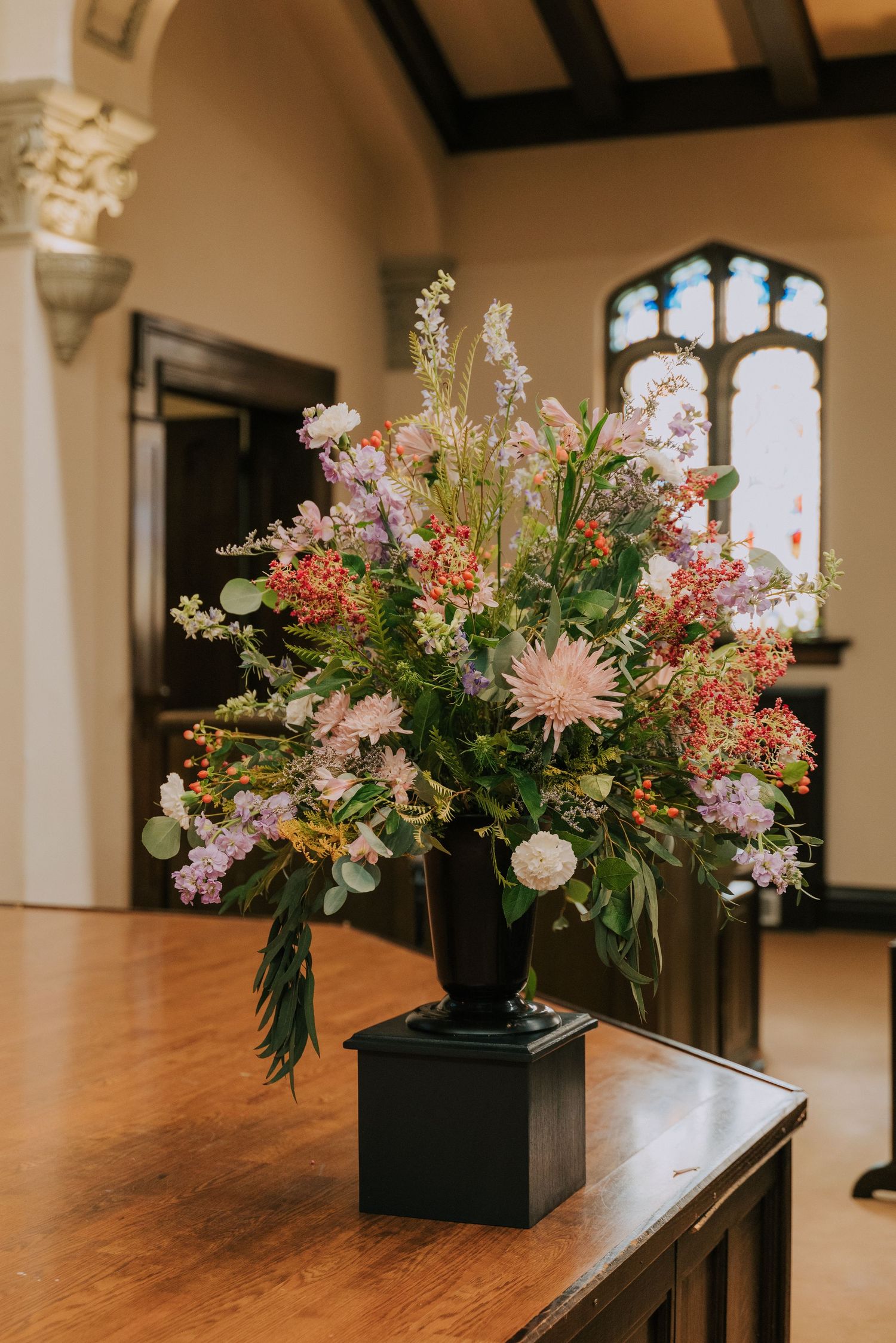 A romantic floral arrangement with pink and peach blooms in a black vase sits atop a wooden table in a grand interior space.