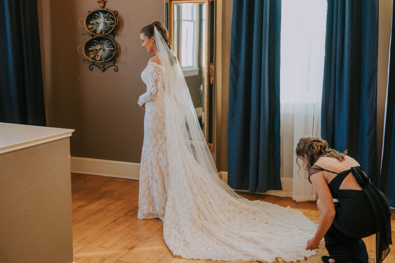 Long white lace wedding gown with flowing cathedral veil being fitted in a room with teal curtains and hardwood floors.