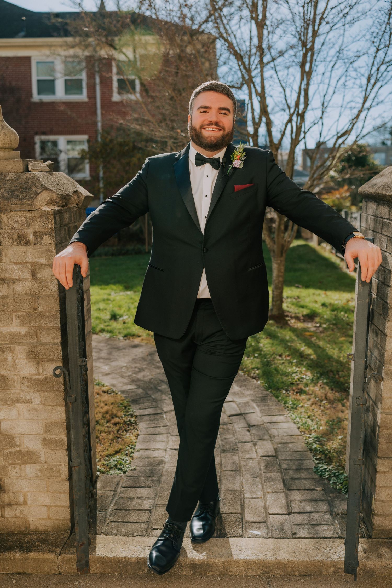 A person in formal black wedding attire poses with arms spread across a stone gate entrance on a brick pathway.