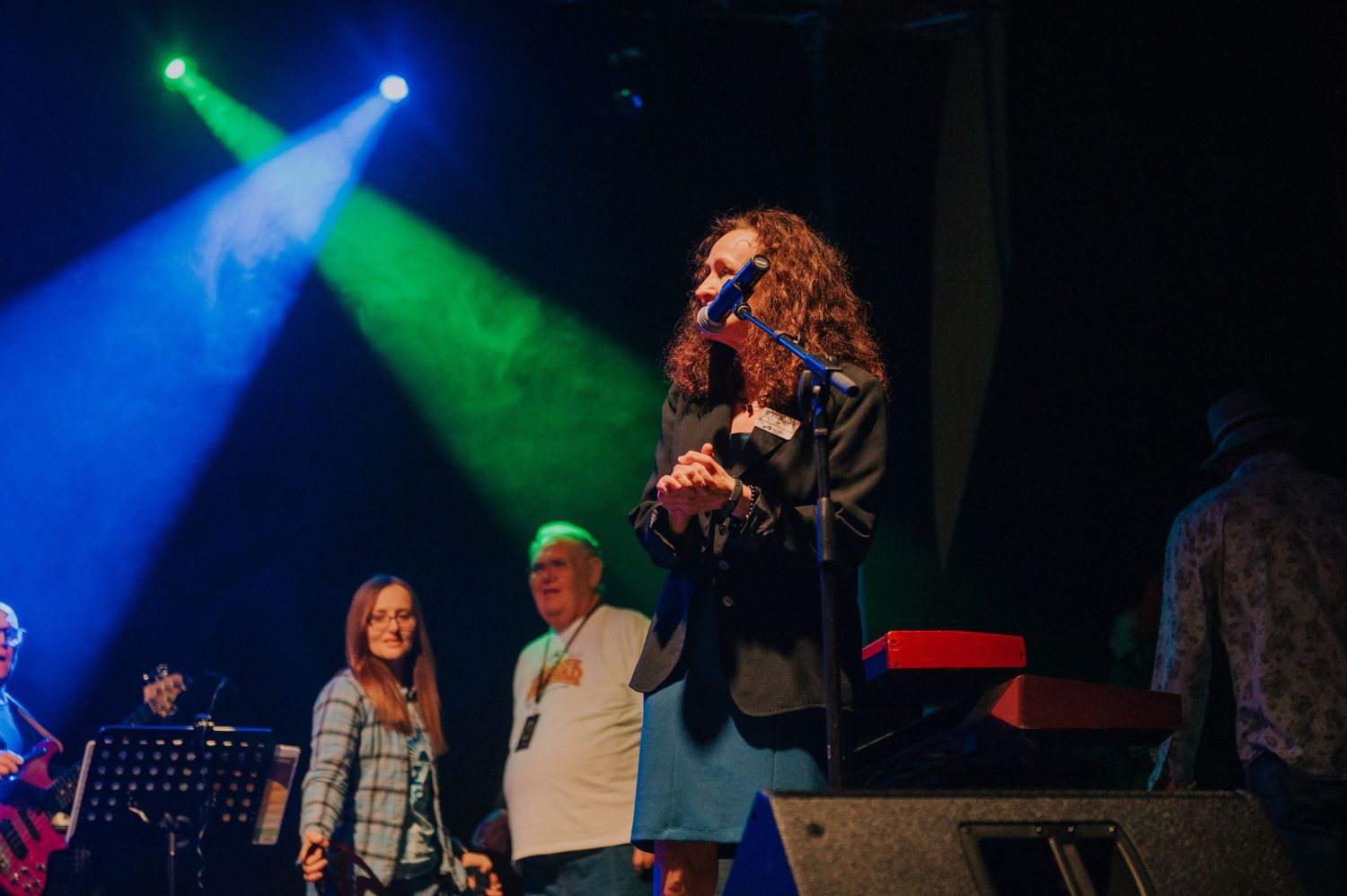Concert performers on stage illuminated by vibrant green and blue spotlights during an energetic musical performance.