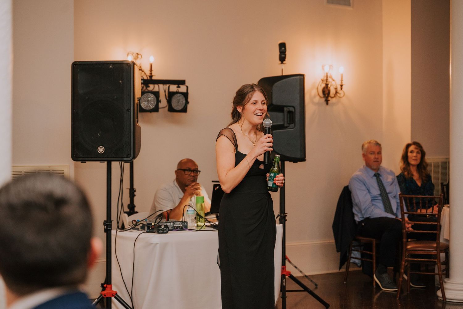 Speaker addressing guests at an indoor event venue with audio equipment and wall sconces.
