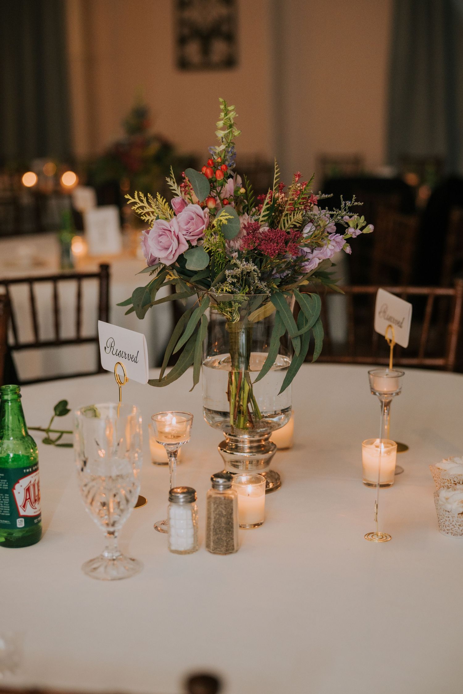 Elegant wedding table setting with pink floral centerpiece, glowing candles, and vintage glassware on white tablecloth.
