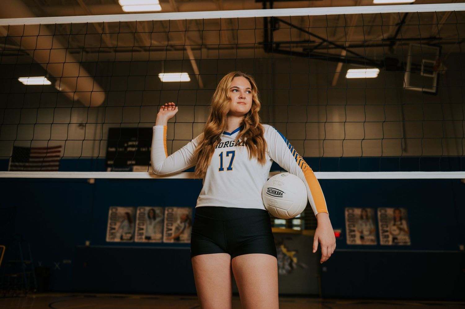 A volleyball player in a white jersey and black shorts stands ready to serve on an indoor court.