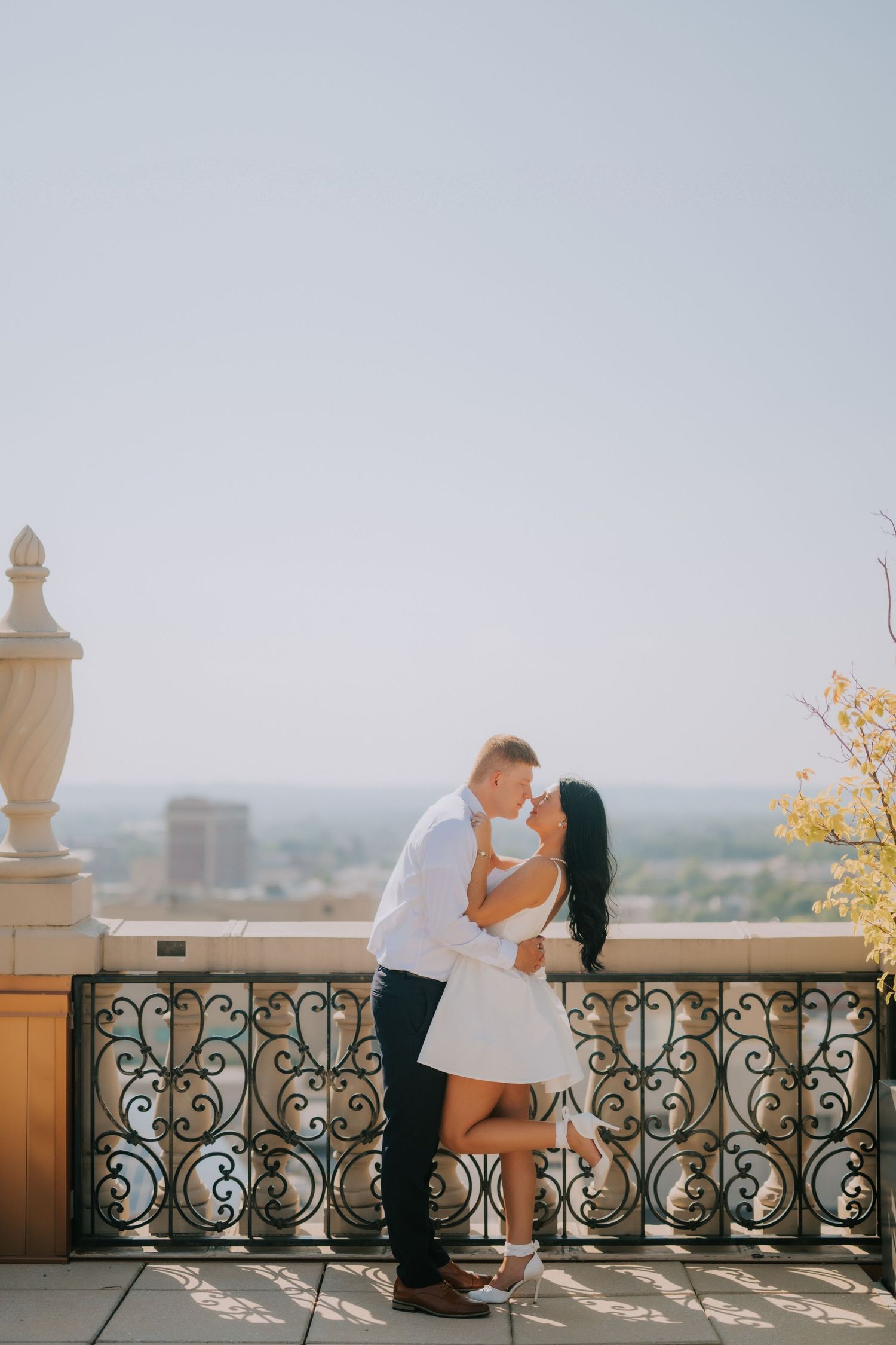 Couple sharing a romantic kiss on an ornate iron balcony with a city skyline backdrop on a sunny day.