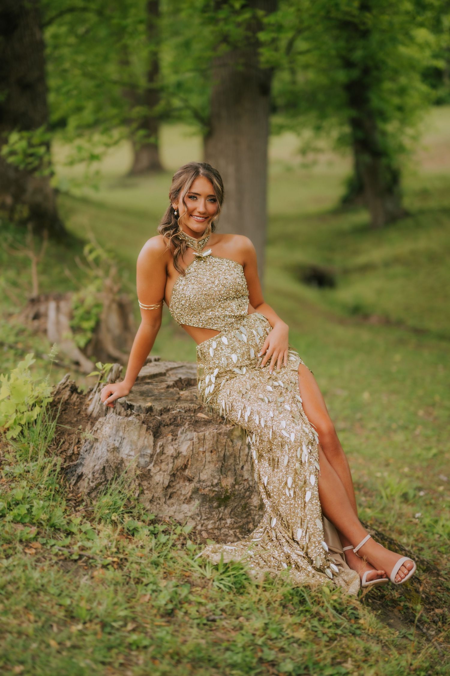 Young woman in gold sequin two-piece formal gown sitting on tree stump in lush green park setting.