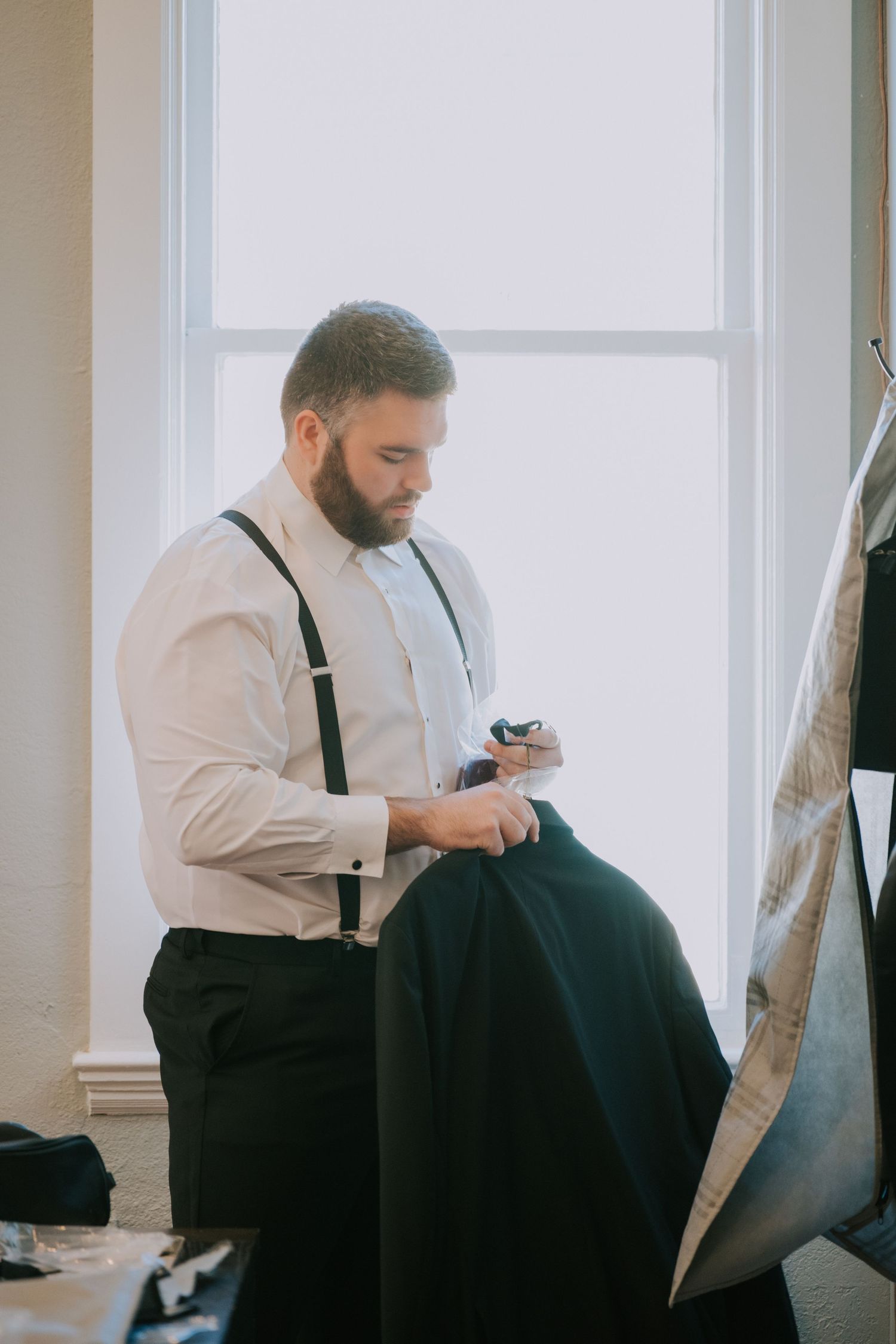 A groomsman in white shirt and suspenders adjusts his wedding attire near a bright window.