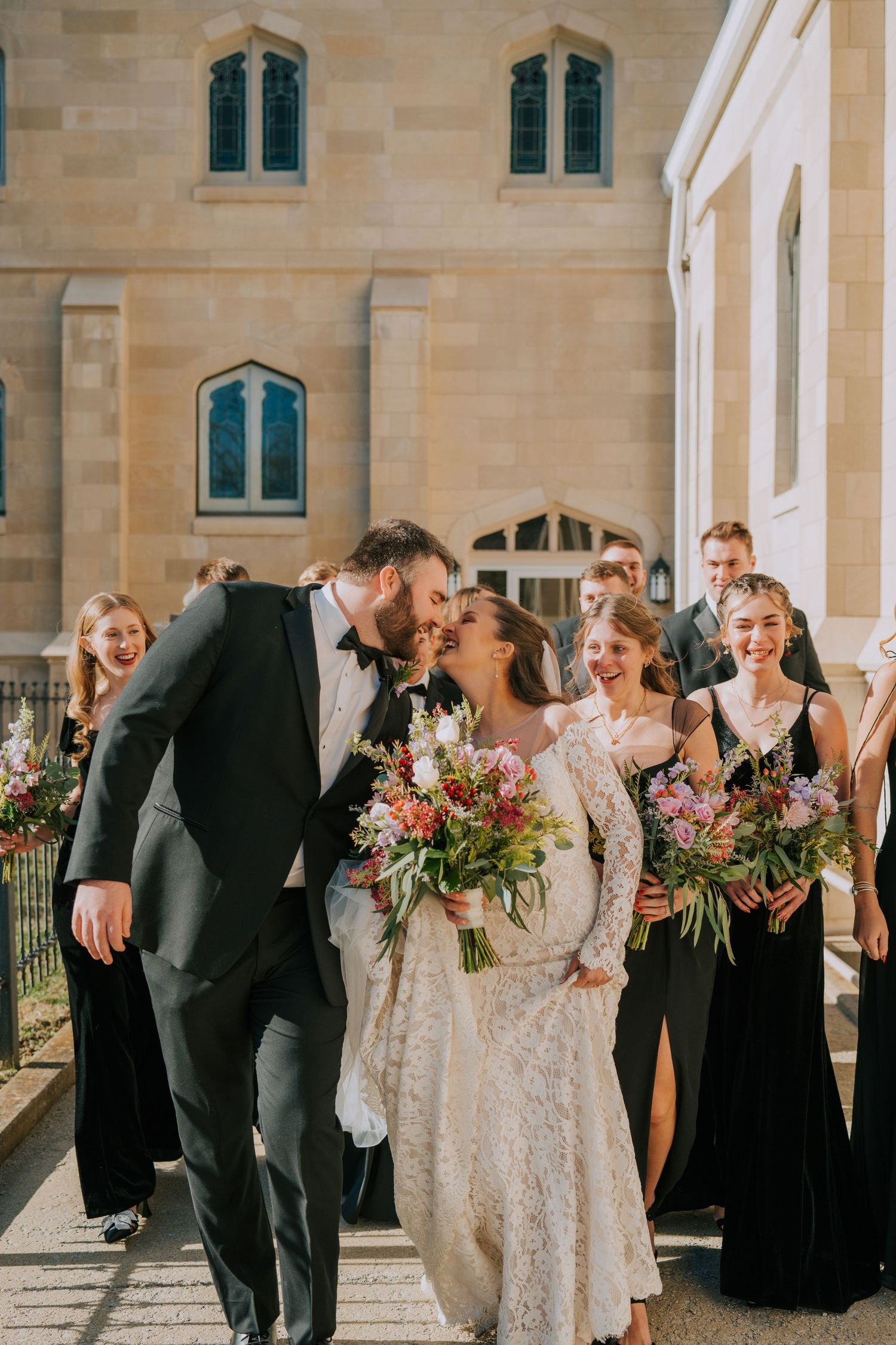 A joyful wedding party shares candid laughter outside a stone church on a sunny day.