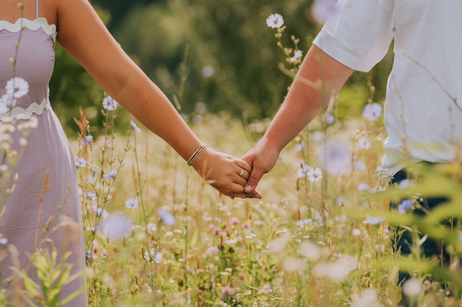 Close-up of two people holding hands among blooming wildflowers in a summer field.