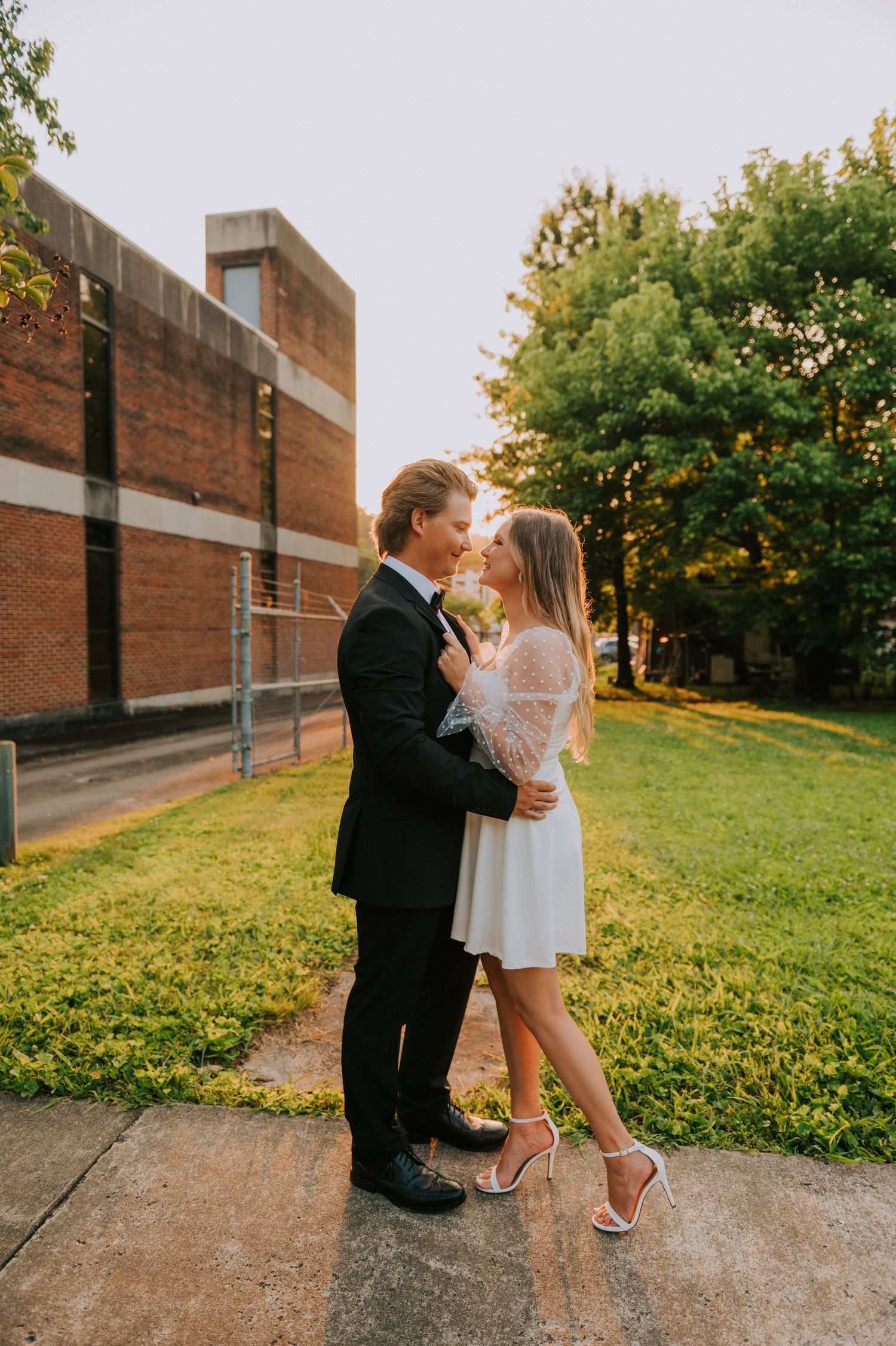 A romantic moment captured on a path surrounded by green grass and brick buildings.