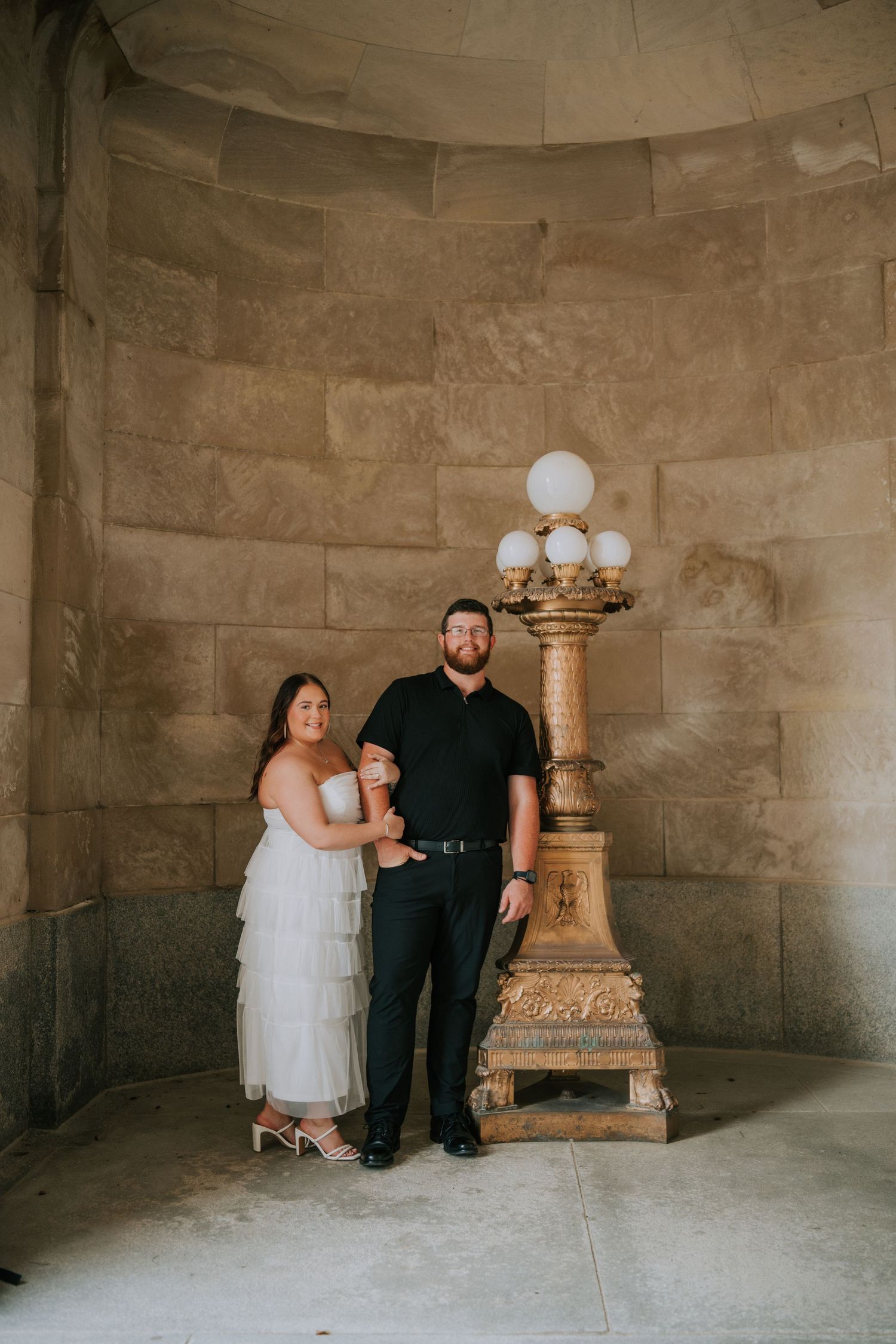 A couple poses in a stone-walled room with vintage light fixtures.
