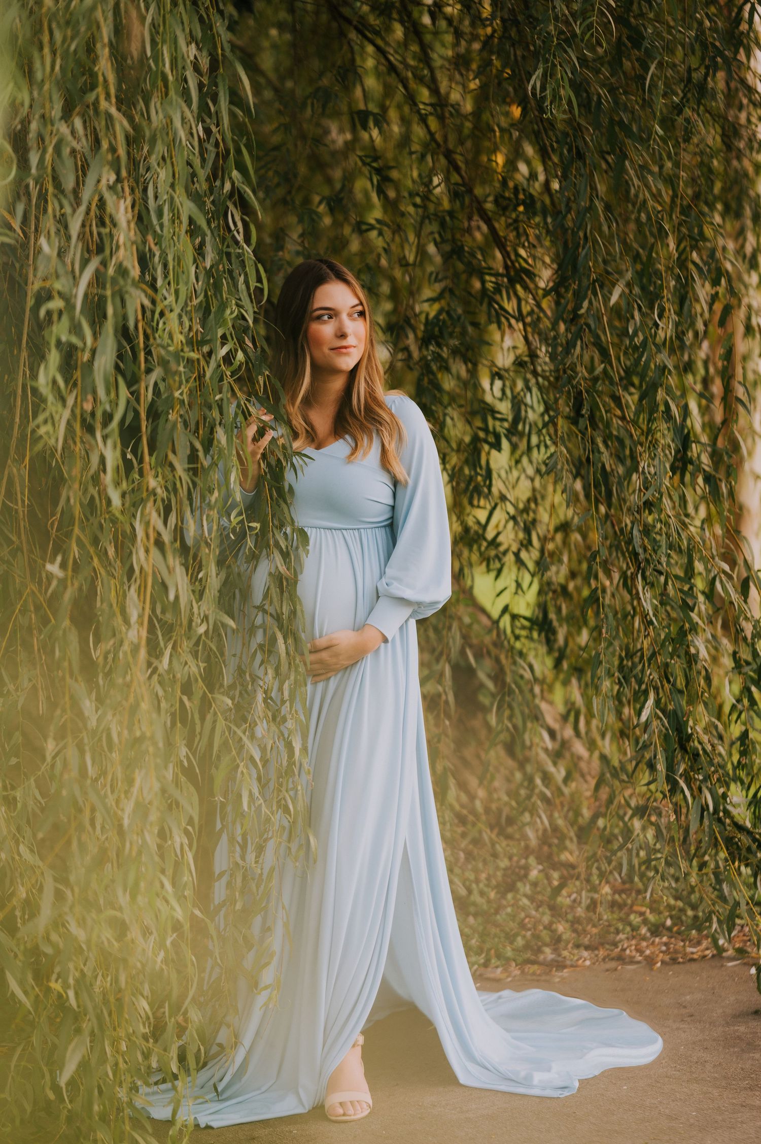A maternity photo session under willow trees with a woman in a flowing light blue dress.