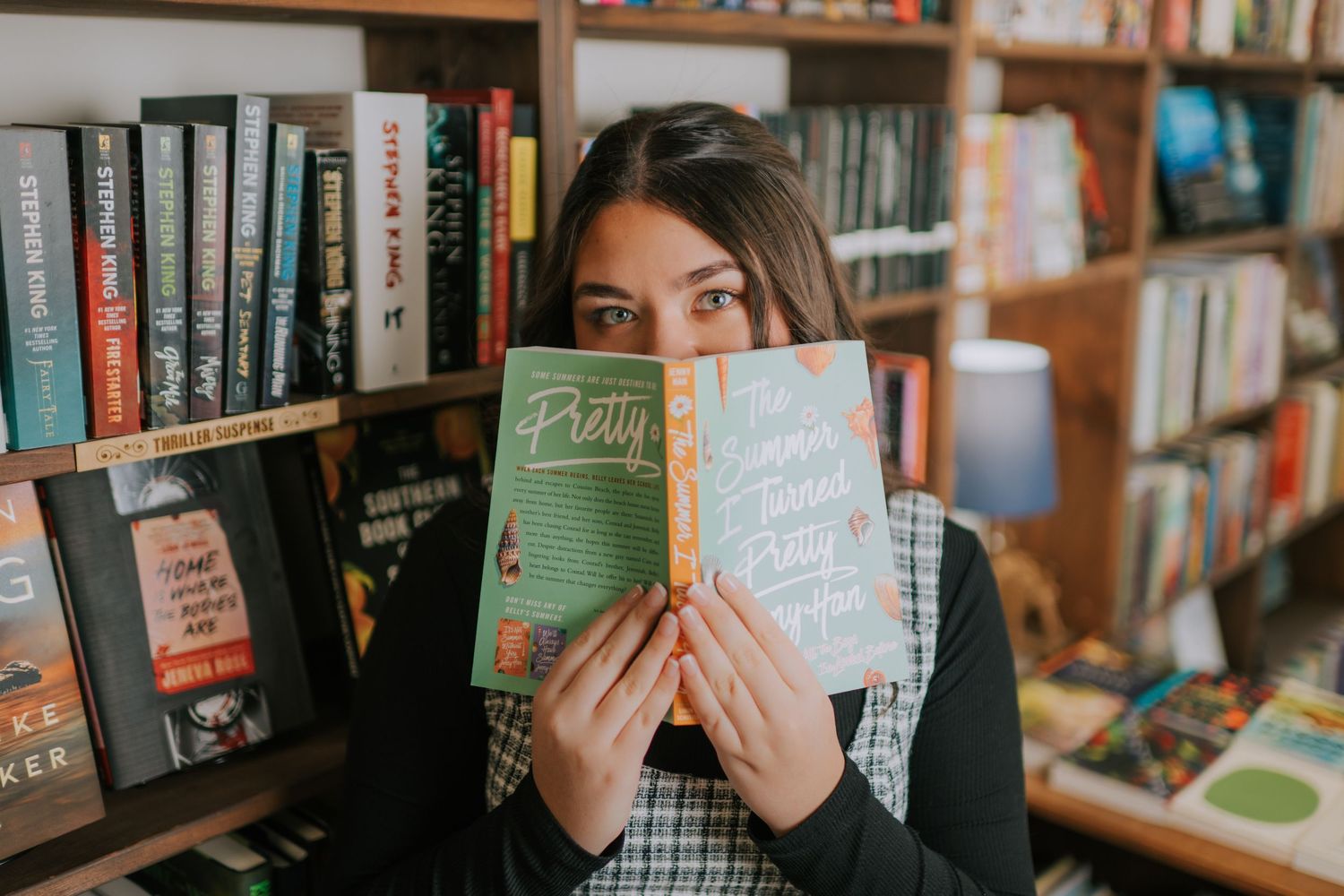 Someone holds up a book in front of their face while standing in a cozy library with wooden bookshelves.