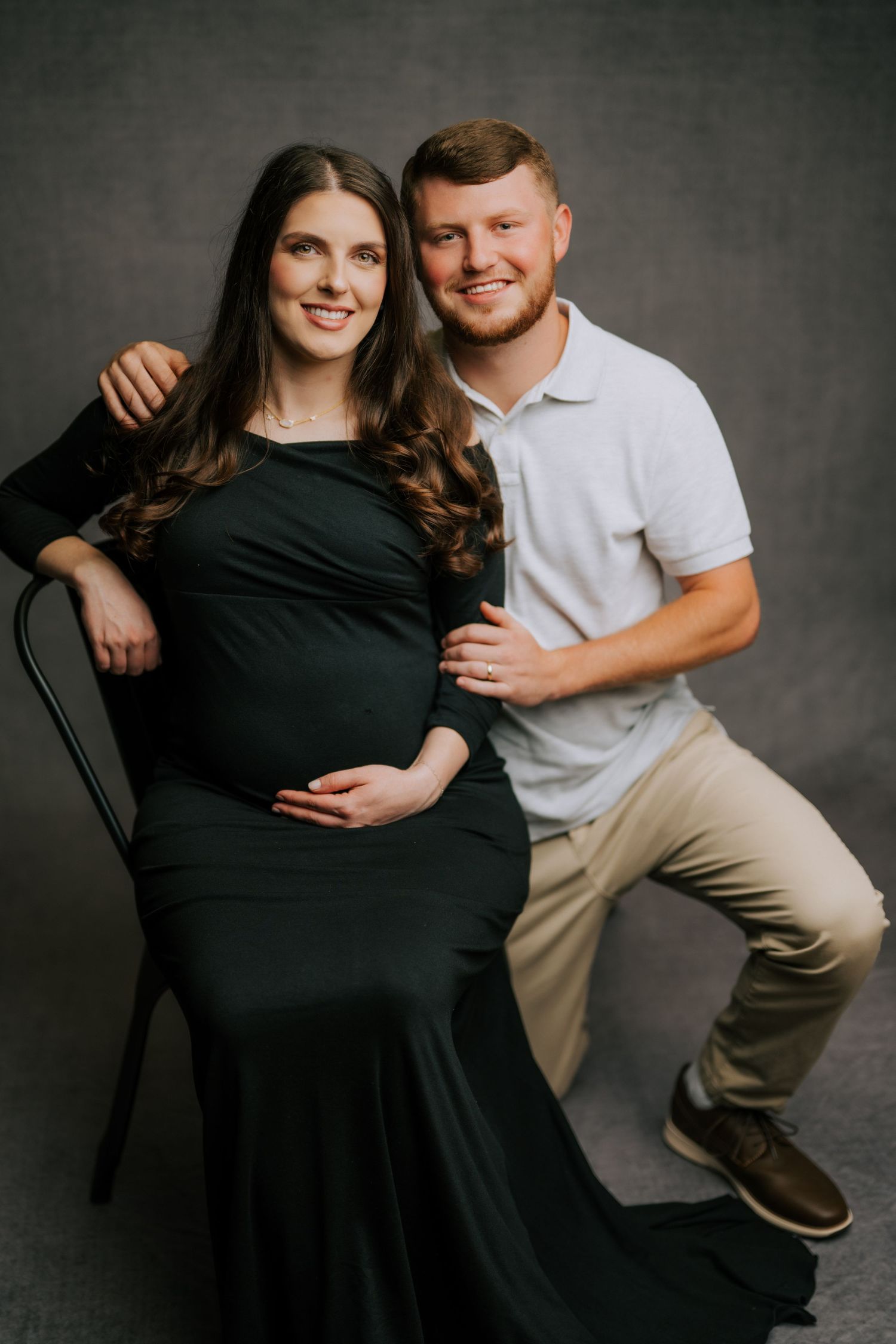 Maternity portrait with couple against dark background, woman in elegant black dress.