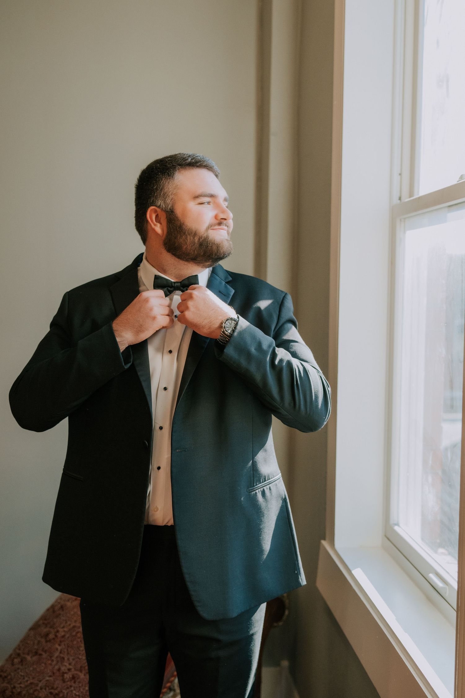 Groom adjusting bowtie while standing by window in classic tuxedo.
