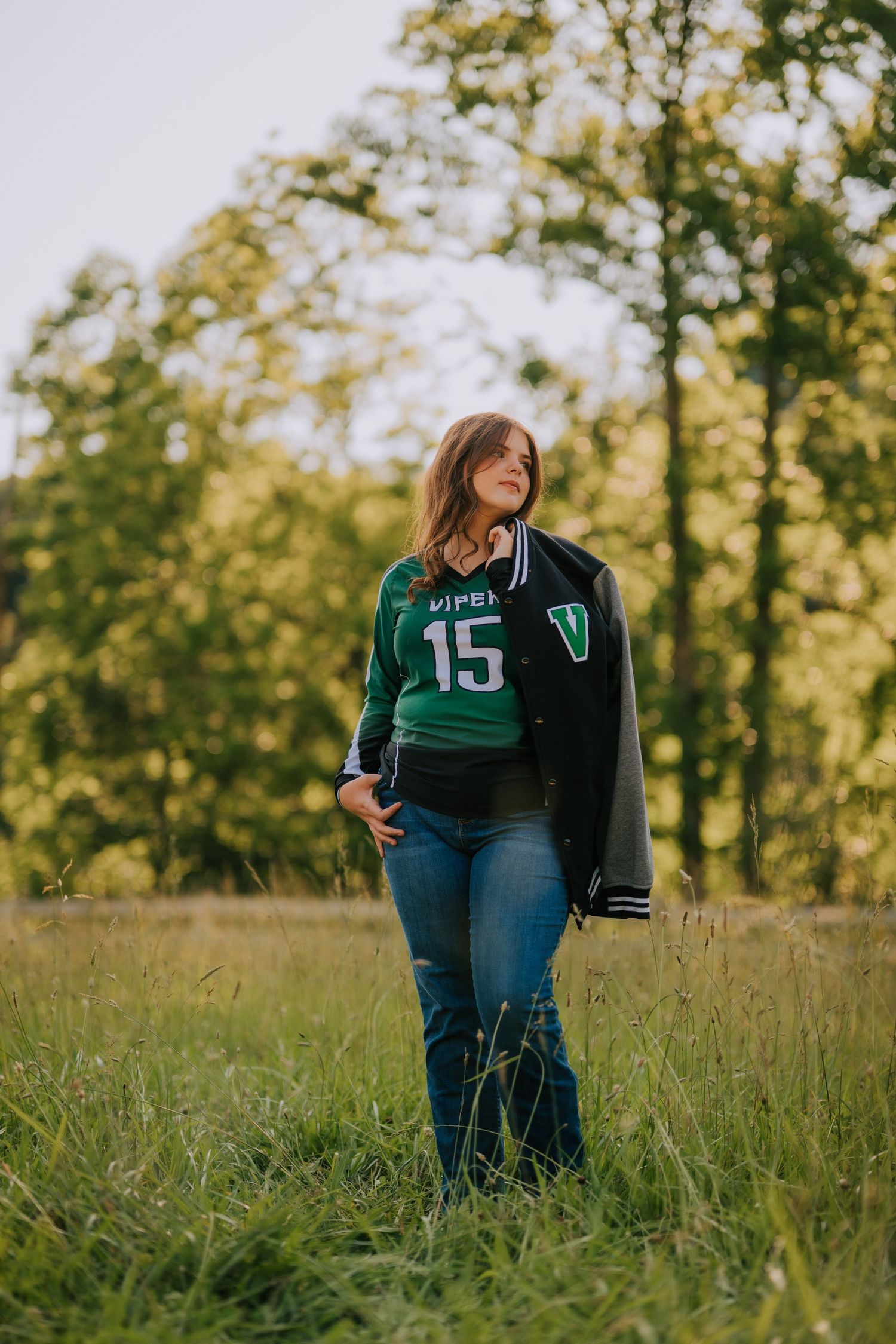 Student wearing green varsity jacket stands in sunny meadow with tall grass.