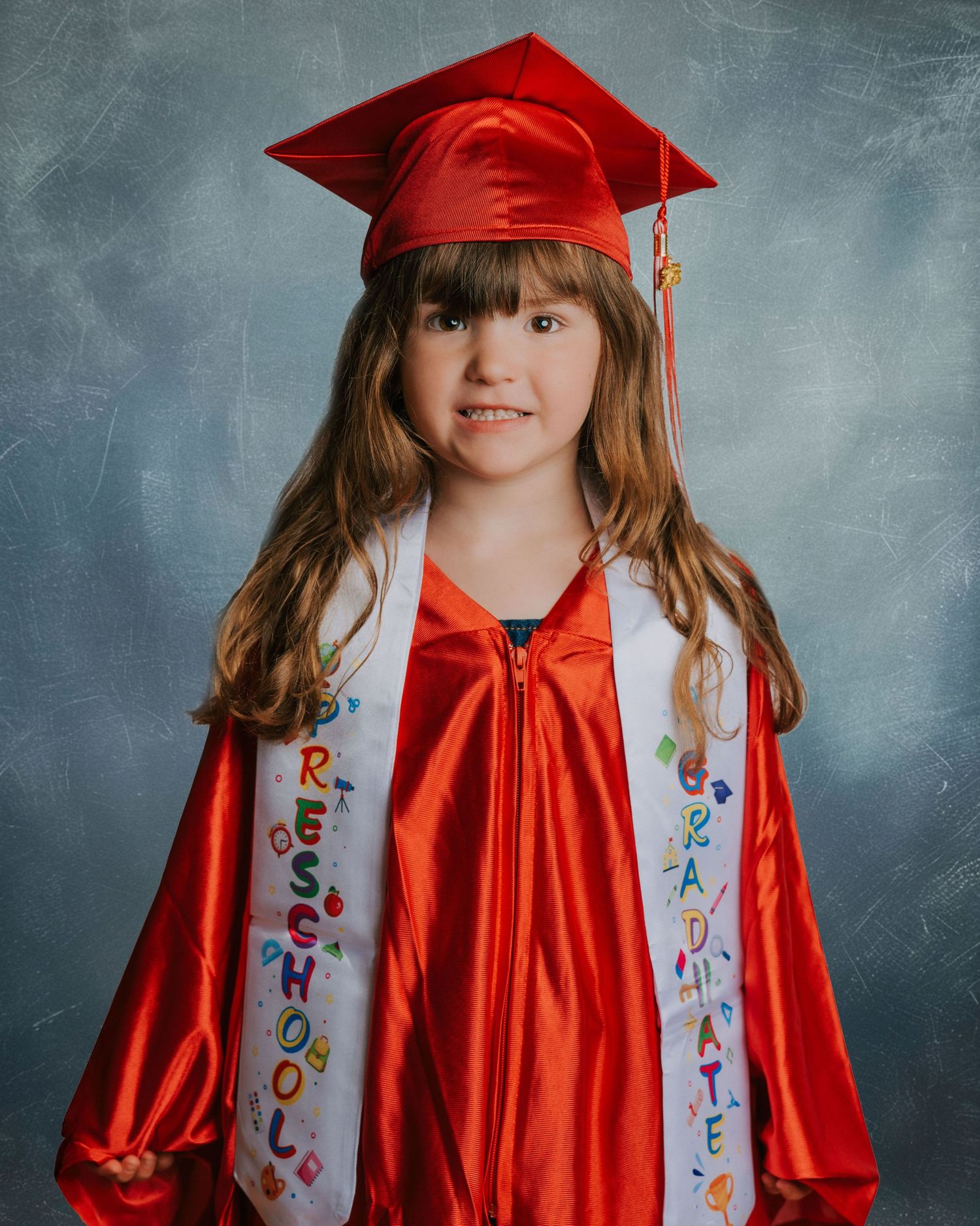 Young girl in red graduation cap and gown wearing a Preschool Graduate sash, smiling for her preschool graduation portrait.