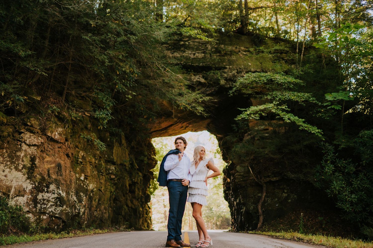 Couple poses together in scenic cave opening surrounded by lush forest landscape.