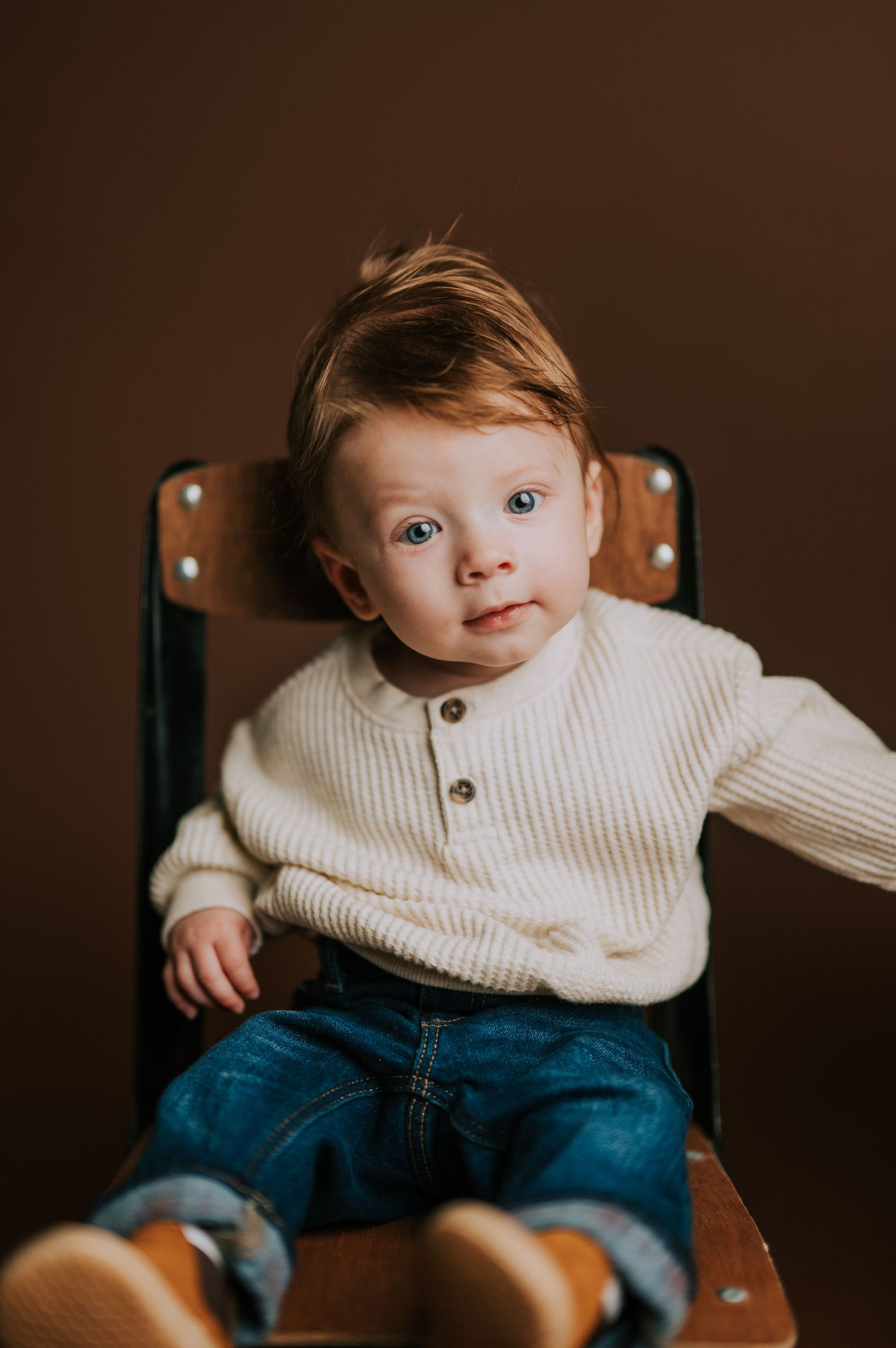 Baby wearing cream sweater and jeans sitting in wooden chair against brown backdrop.