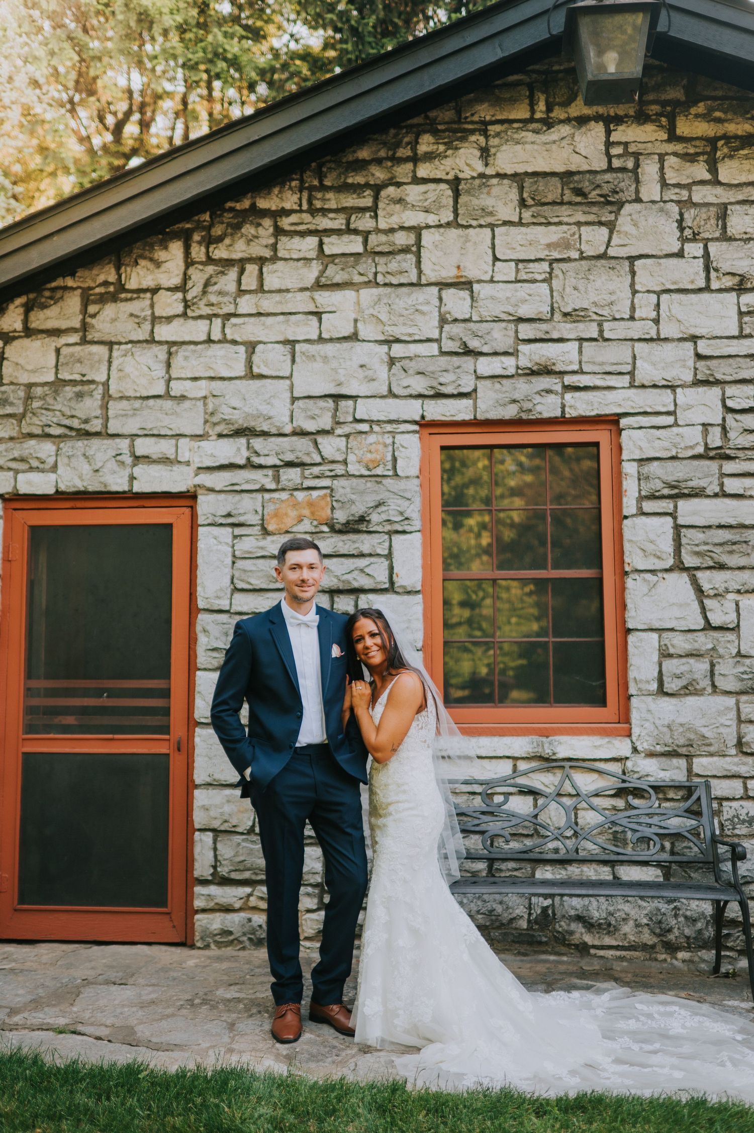 Wedding couple standing against stone wall of rustic building with red-trimmed windows.