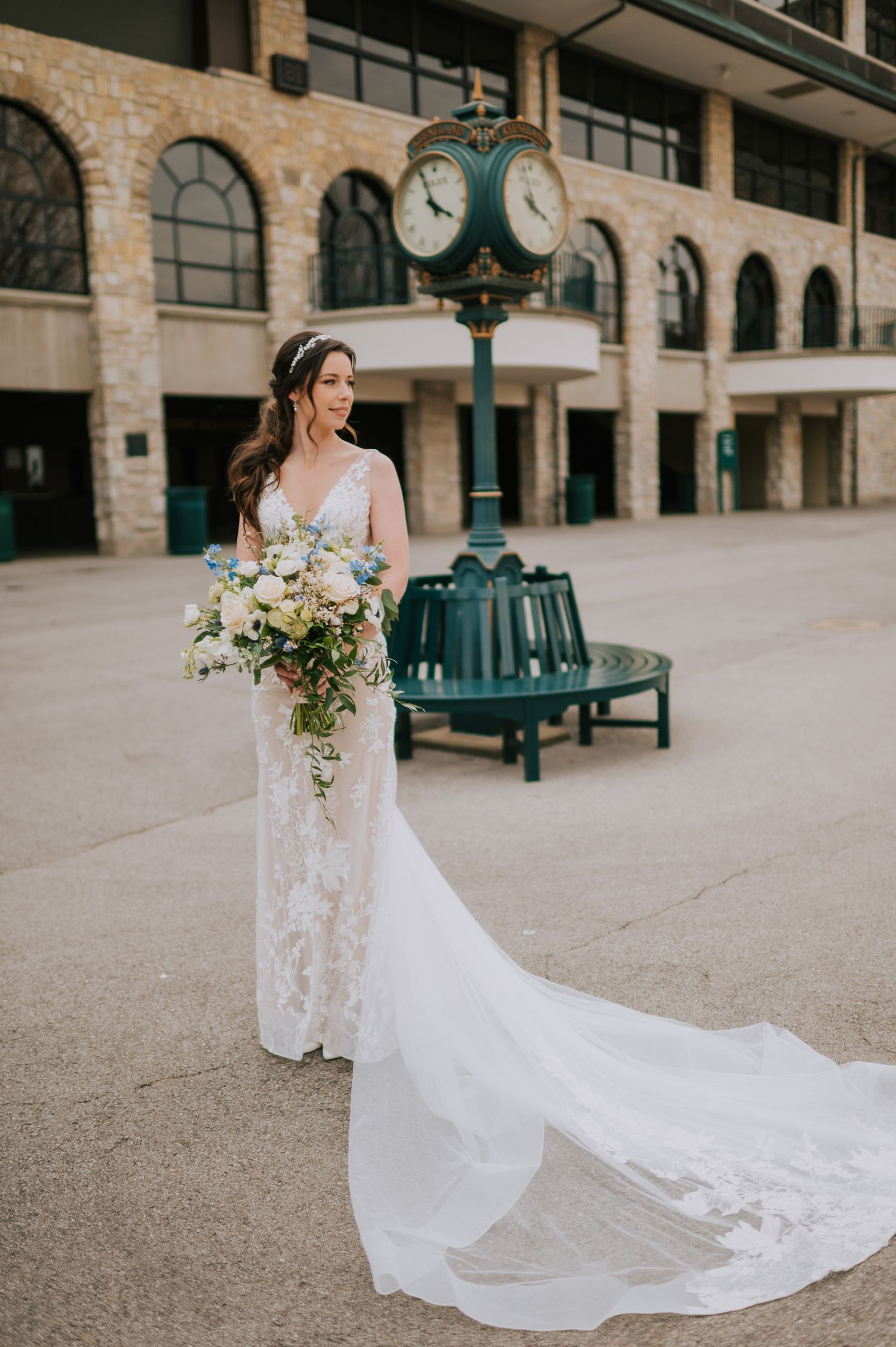 Bride in flowing lace gown with long train standing near vintage clock tower.