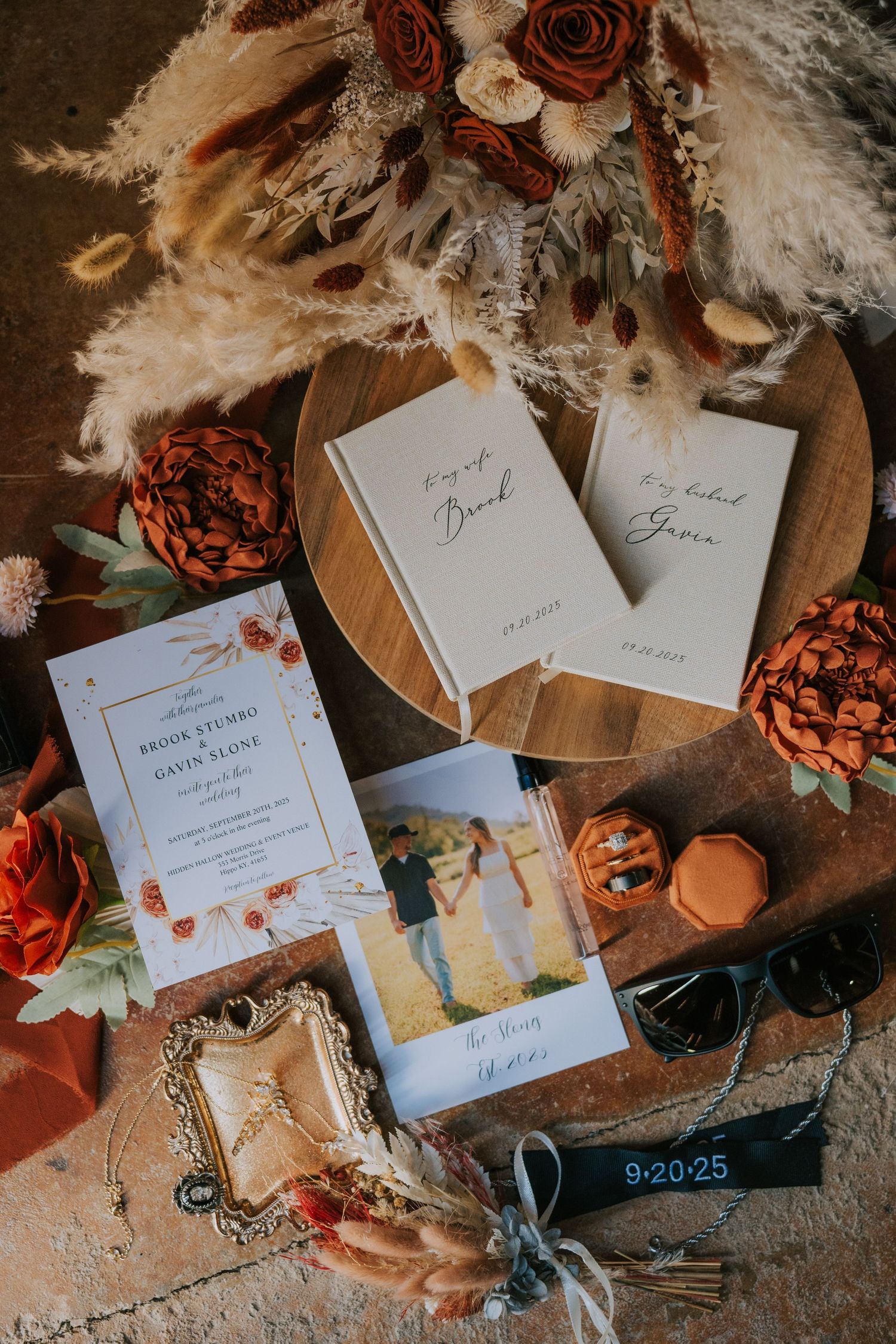Wedding stationery flatlay with dried roses, invitation cards, vow books and decorative details on rustic wooden surface.