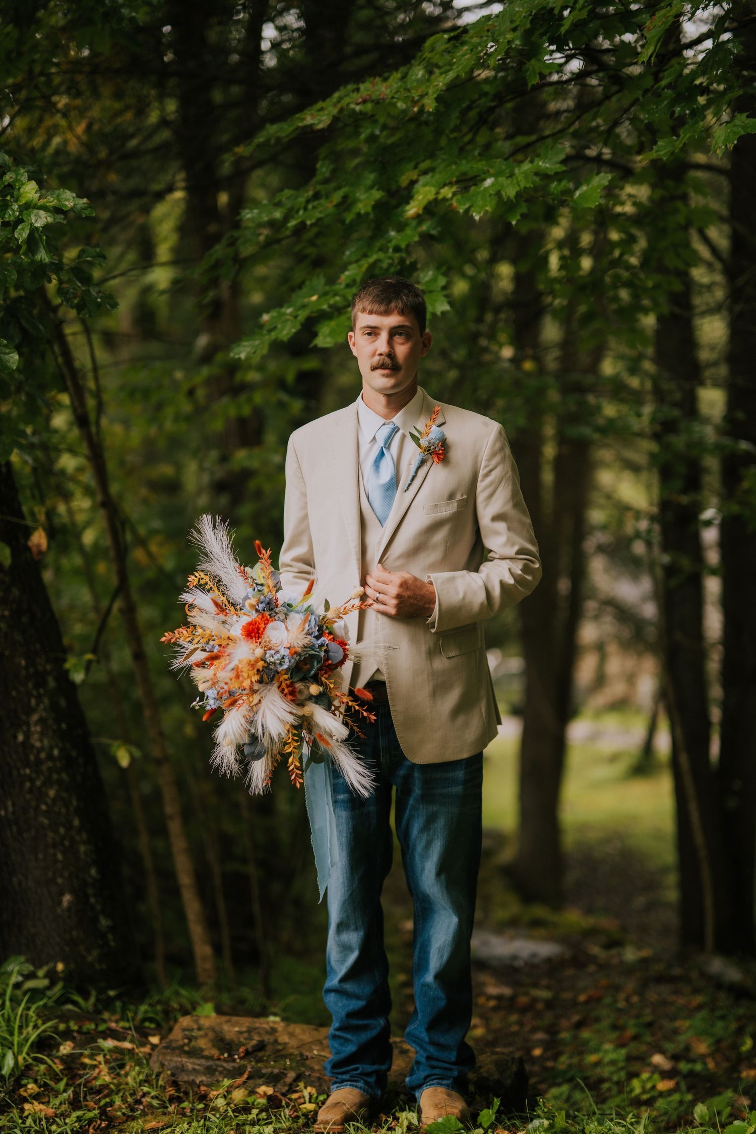 A casual wedding outfit combining beige jacket and jeans in an outdoor woodland setting.