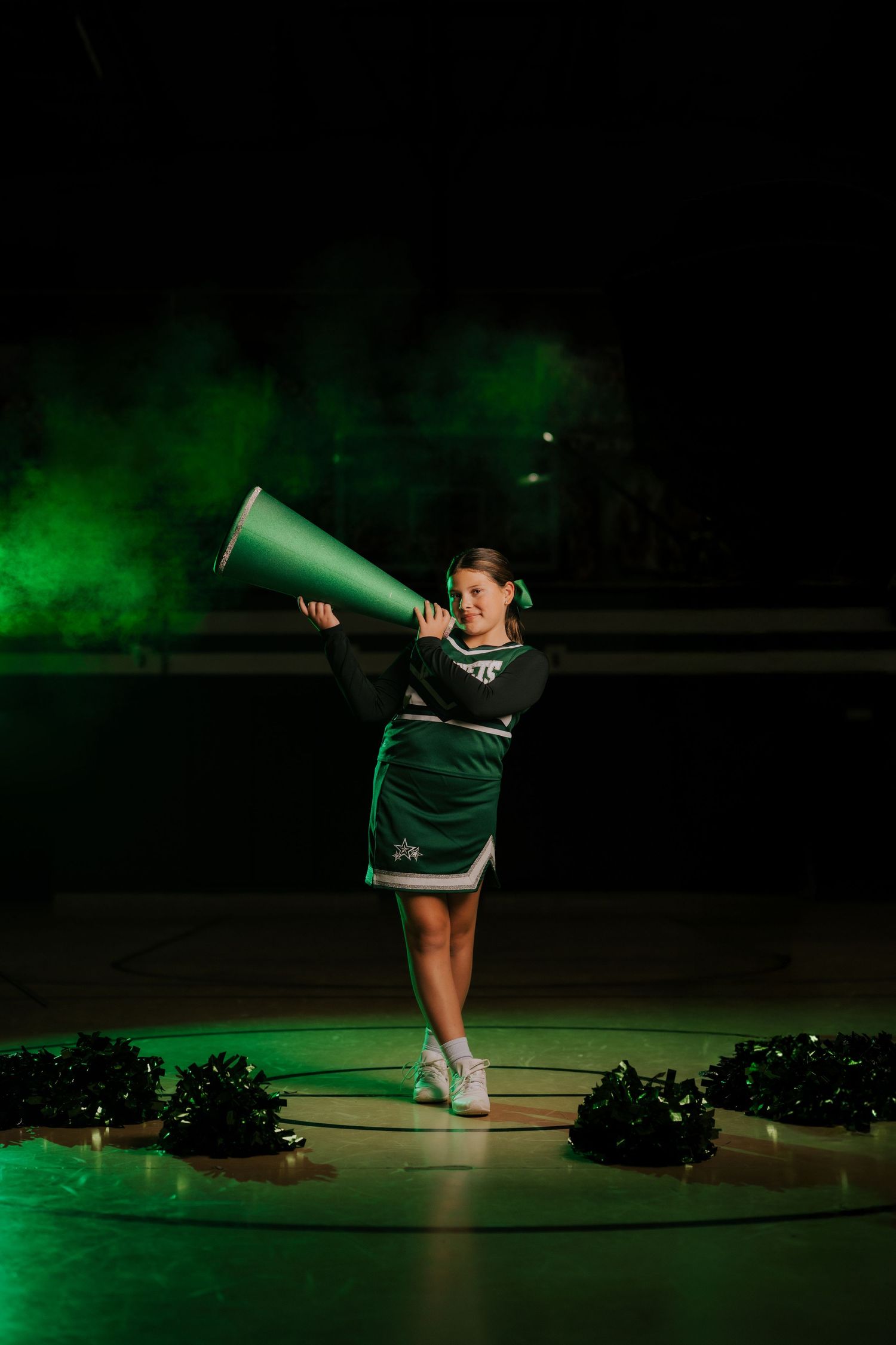 Cheerleader performs with megaphone under dramatic green spotlight on dark stage.