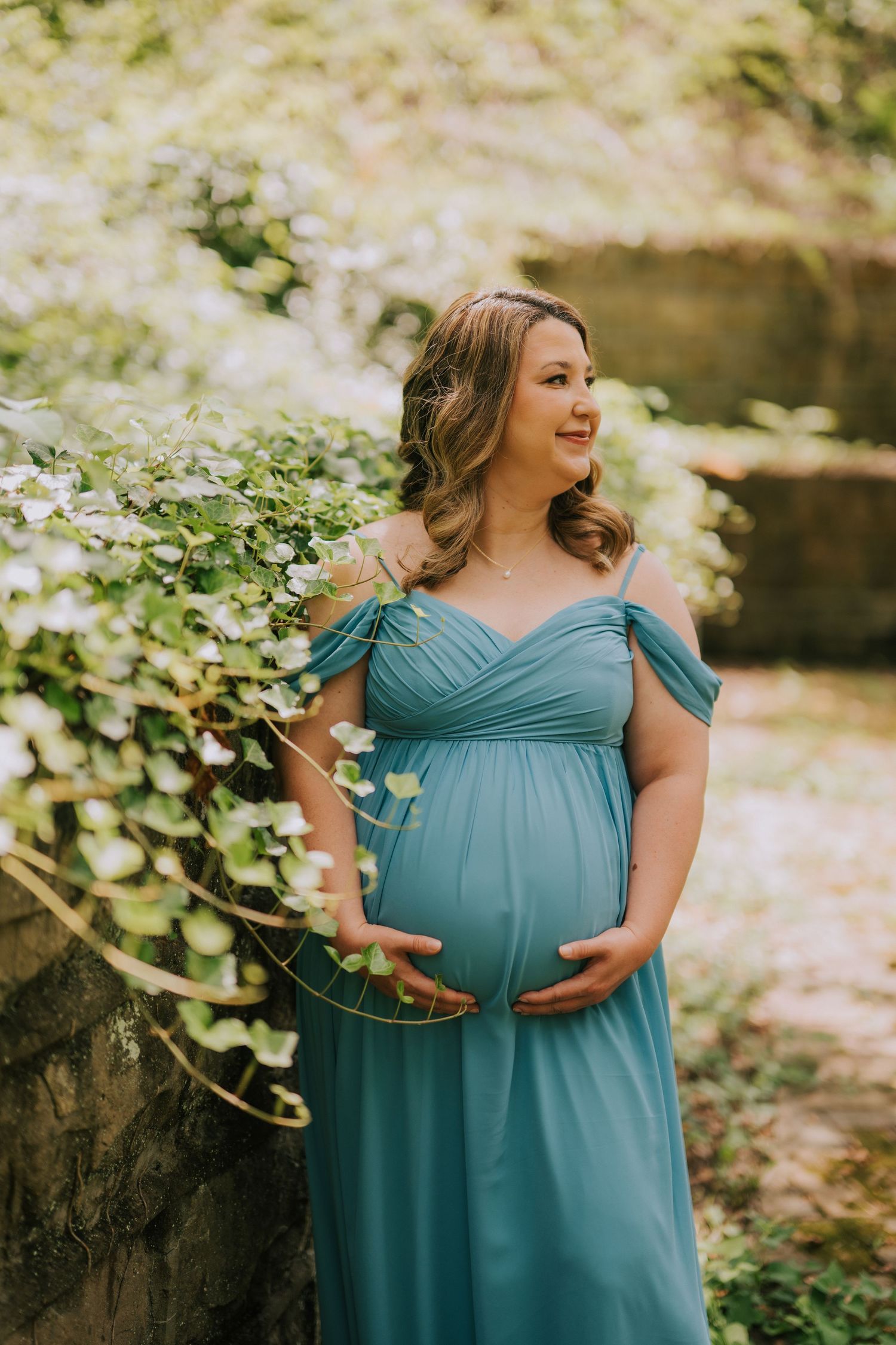 Expectant mother in teal dress poses gracefully among blooming foliage.
